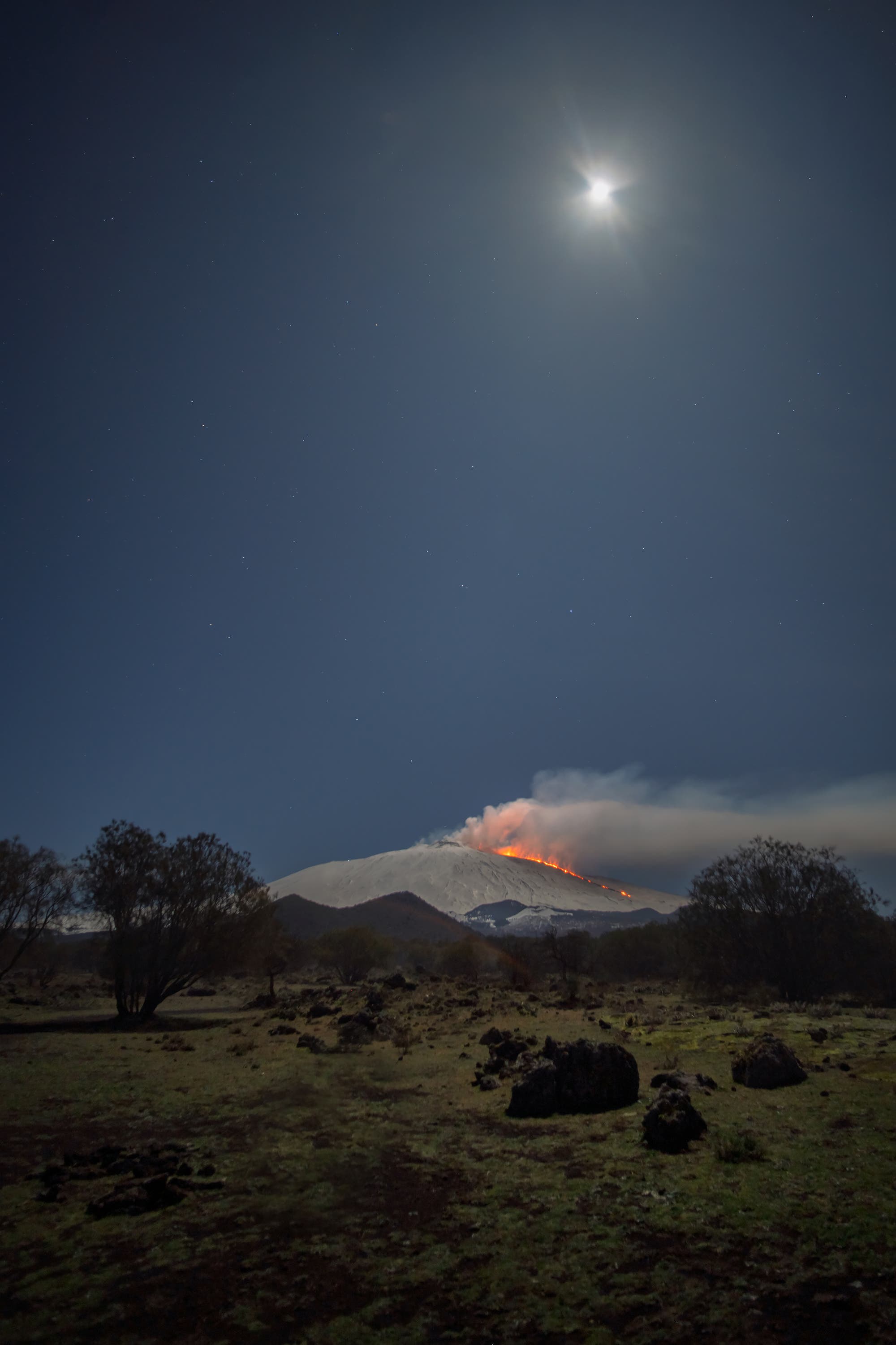 The moon above the erupting Etna volcano