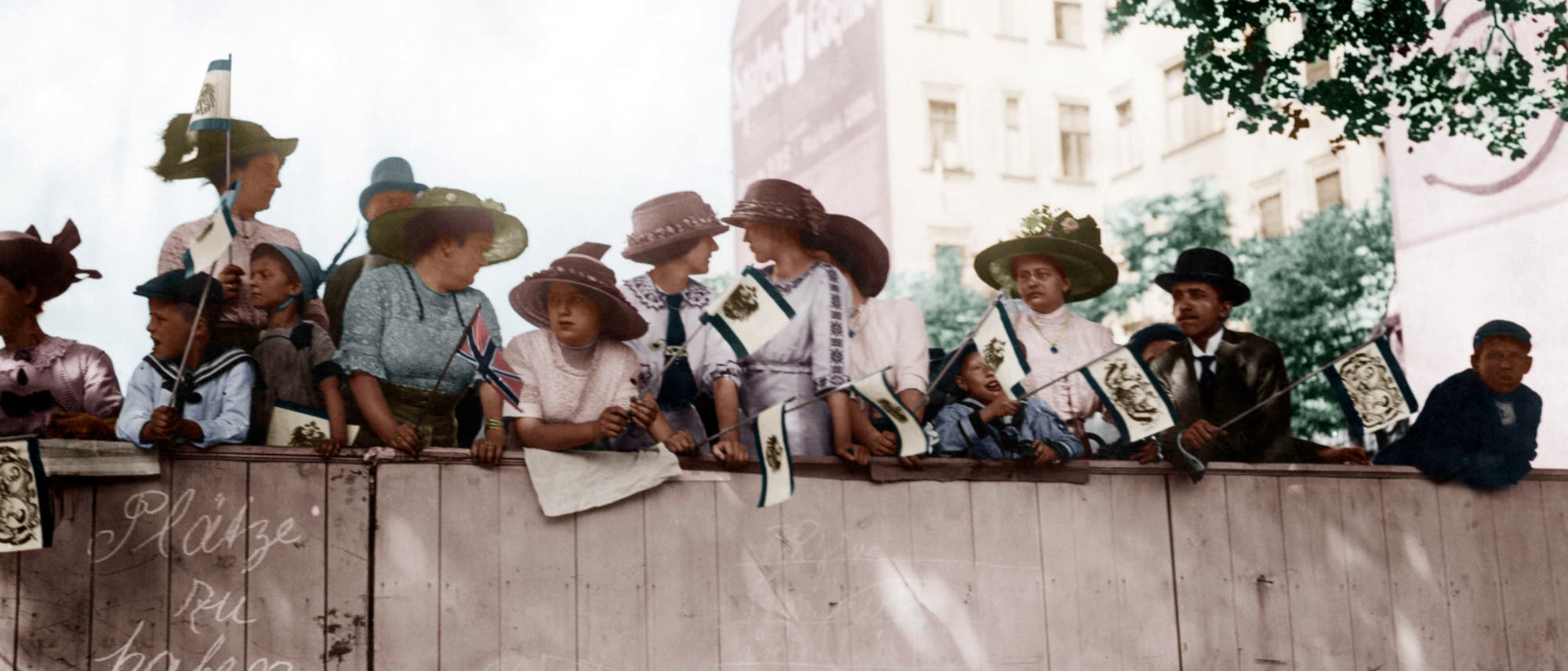 Frühjahrsparade in Berlin, das digital kolorierte Foto von Otto Haeckel aus dem Jahr 1910 zeigt Frauen, die auf einen Bauzaun geklettert sind, um eine bessere Sicht zu haben Frühjahrsparade in Berlin, das digital kolorierte Foto von Otto Haeckel aus dem Jahr 1910 zeigt Frauen, die auf einen Bauzaun geklettert sind, um eine bessere Sicht zu haben