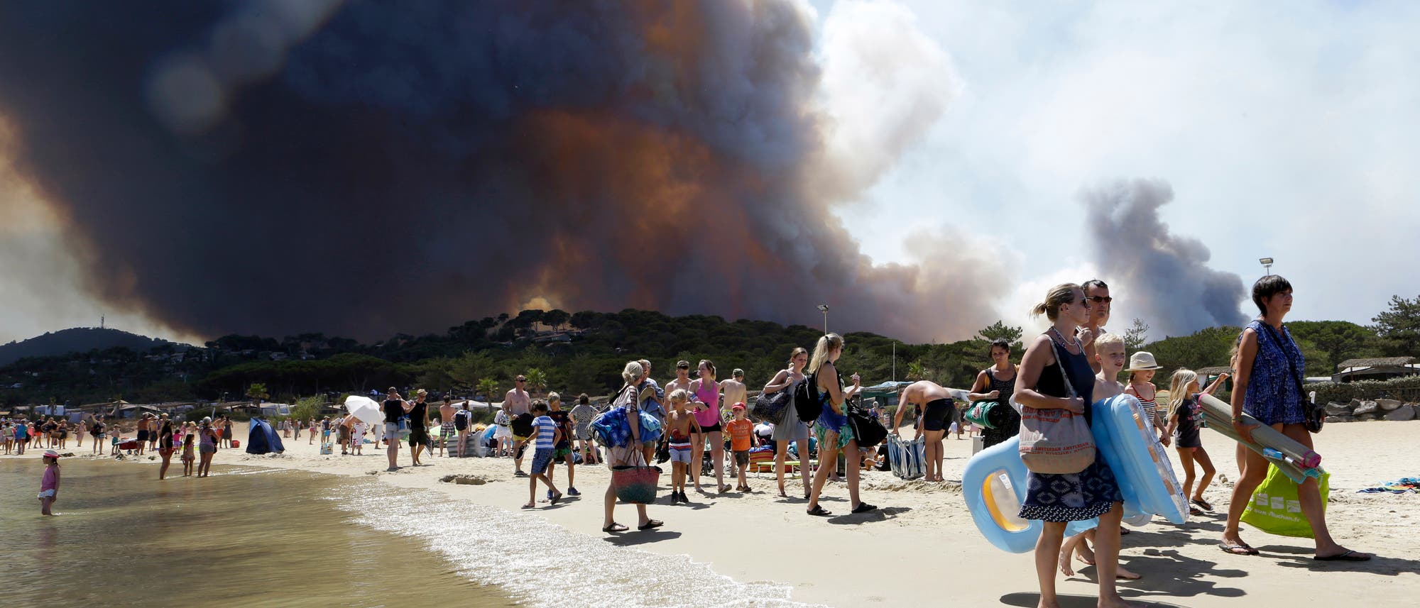 Ein Strand wird evakuiert, während hinter den Dünen im Hintergrund bereits dunkler Rauch eines Waldbrands aufsteigt. Ein Strand wird evakuiert, während hinter den Dünen im Hintergrund bereits dunkler Rauch eines Waldbrands aufsteigt.