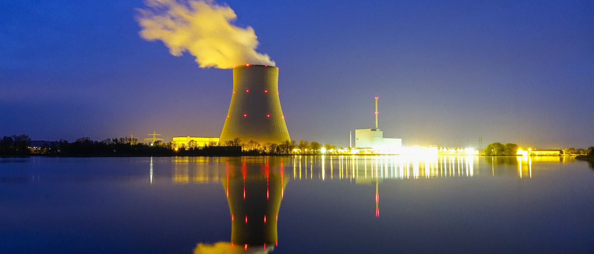Kühlturm mit Dampfwolke und hell erleuchtete Kraftwerksgebäude in der Dämmerung, fotografiert vom gegenüberliegenden Flussufer. Kühlturm mit Dampfwolke und hell erleuchtete Kraftwerksgebäude in der Dämmerung, fotografiert vom gegenüberliegenden Flussufer.