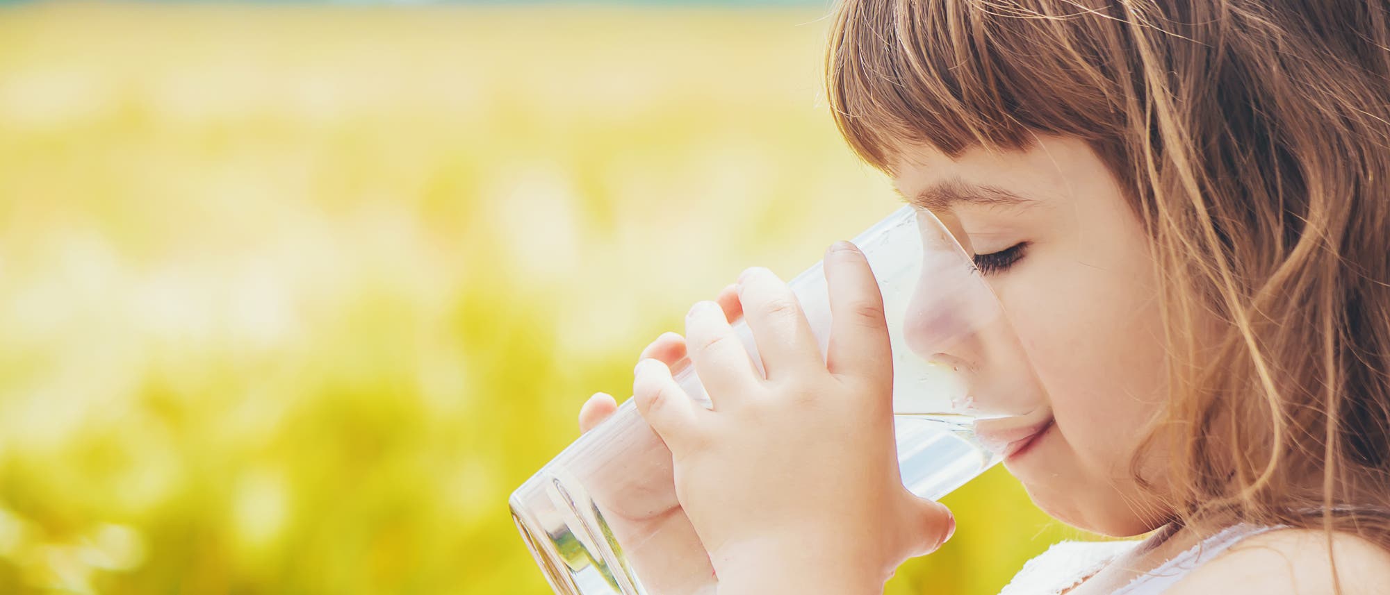 Ein Mädchen trinkt Wasser aus einem Glas. Im Hintergrund unscharf ein Rapsfeld. Ein Mädchen trinkt Wasser aus einem Glas. Im Hintergrund unscharf ein Rapsfeld.