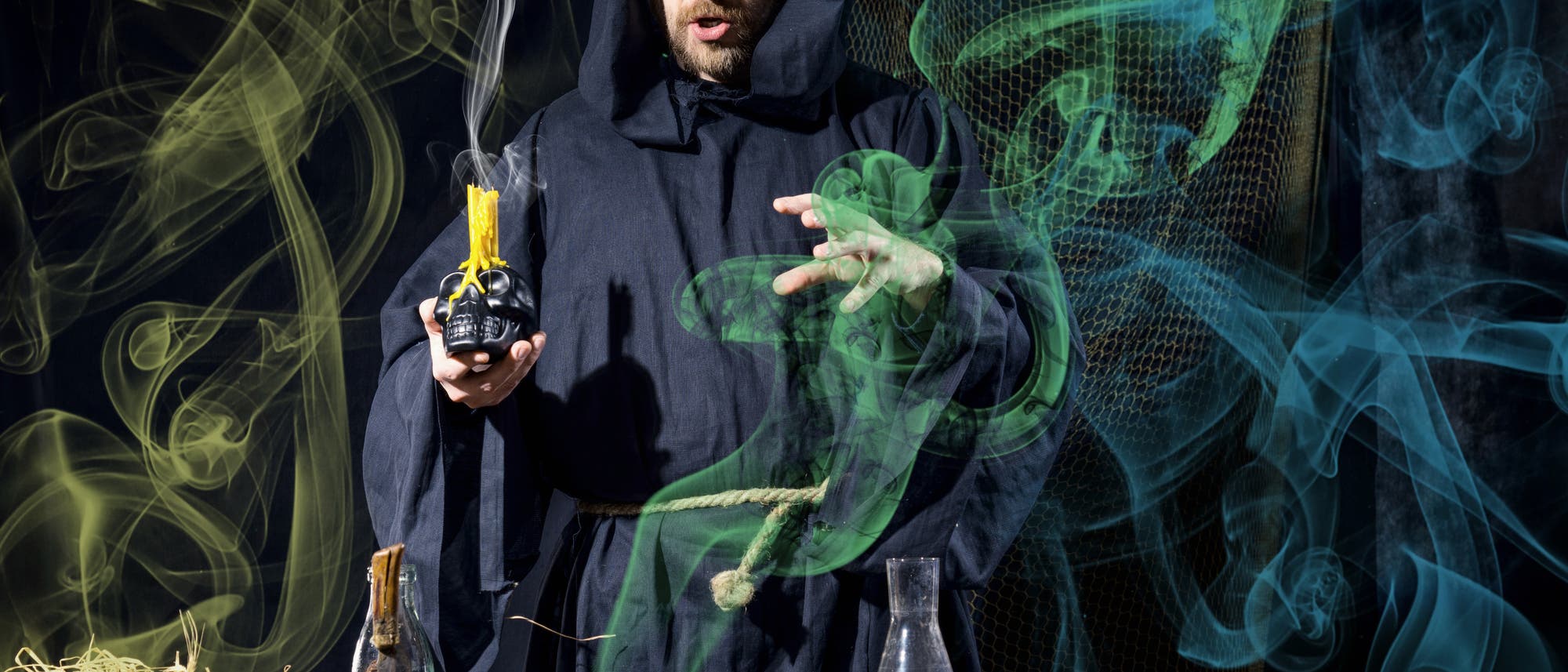Ein Alchemist mit Kutte hält eine Kerze auf einem Totenschädel und forscht im bunten Dunst Ein Alchemist mit Kutte hält eine Kerze auf einem Totenschädel und forscht im bunten Dunst