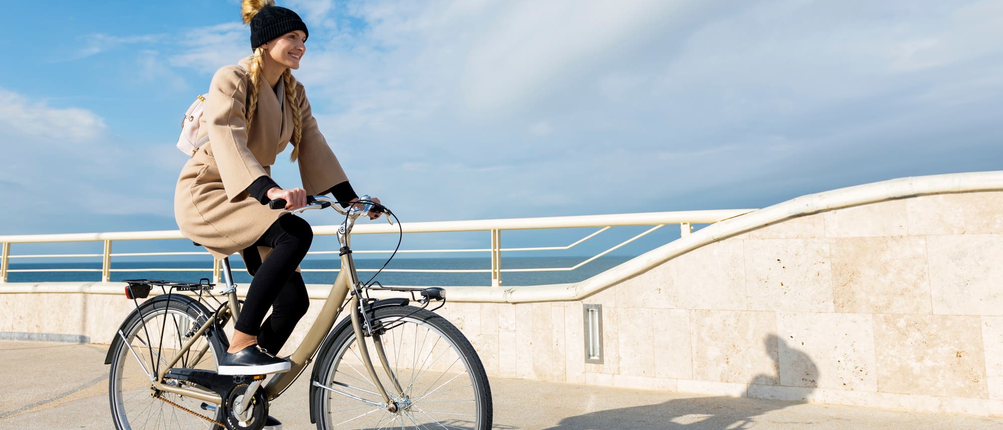 Eine Frau in einem braunen Mantel fährt an einer Strandpromenade Fahrrad. Eine Frau in einem braunen Mantel fährt an einer Strandpromenade Fahrrad.