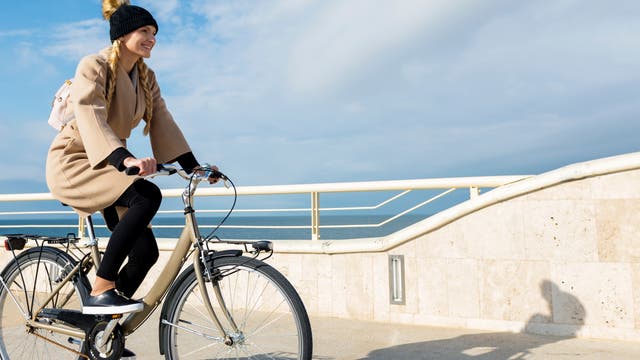Eine Frau in einem braunen Mantel fährt an einer Strandpromenade Fahrrad.