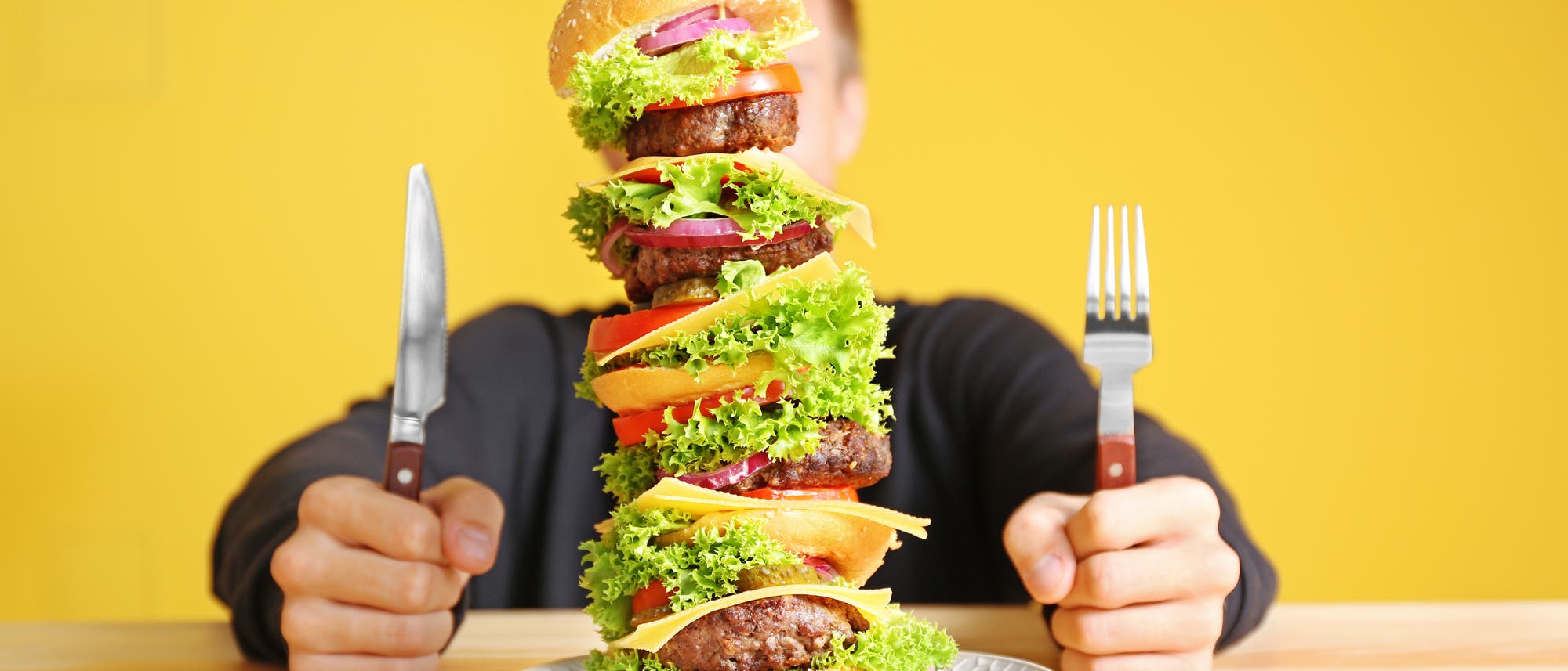 Ein junger Mann, dessen Gesicht hinter dem Megaburger vor ihm auf dem Teller verschwindet Ein junger Mann, dessen Gesicht hinter dem Megaburger vor ihm auf dem Teller verschwindet