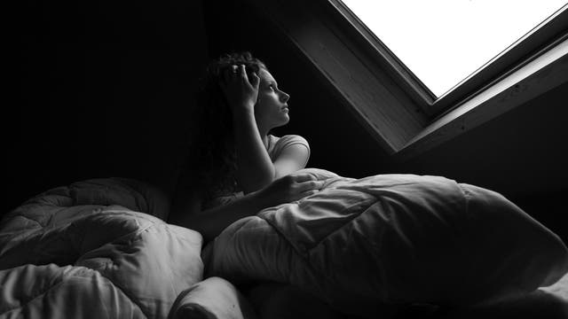 Eine Frau liegt im Bett und sieht durch ein Dachfenster in den grauen Himmel.