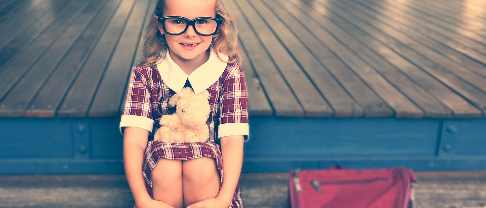 Ein junges Mädchen mit großer Brille sitzt mit angezogenen Beinen auf einer Treppe. Neben ihr steht eine rote Schultasche. Ein junges Mädchen mit großer Brille sitzt mit angezogenen Beinen auf einer Treppe. Neben ihr steht eine rote Schultasche.
