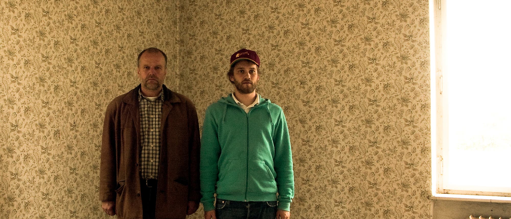 Vater und Sohn stehen in einem leeren Wohnraum, der von einer auffälligen alten Tapete geziert wird. Vater und Sohn stehen in einem leeren Wohnraum, der von einer auffälligen alten Tapete geziert wird.