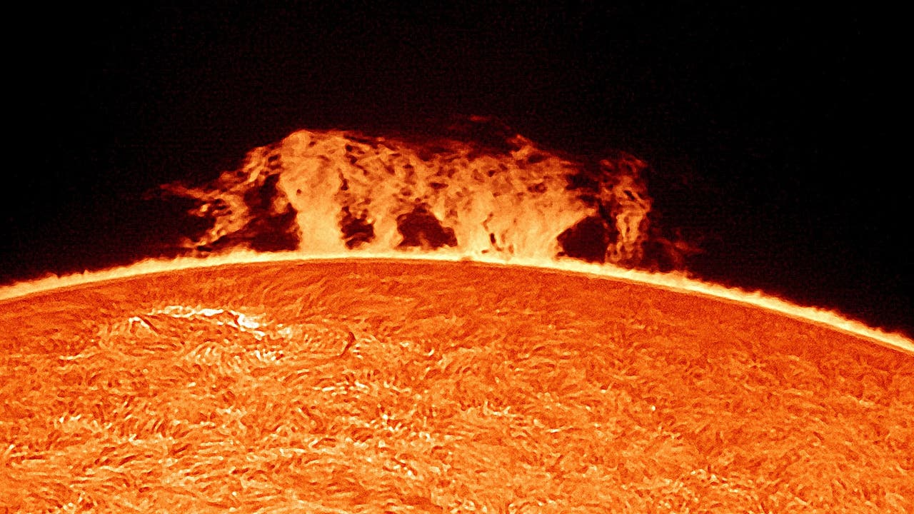 Die Abbildung zeigt eine Nahaufnahme der Sonnenoberfläche mit einer beeindruckenden Sonneneruption, die als leuchtende, flammenartige Struktur über dem Rand der Sonne sichtbar ist. Die Oberfläche der Sonne erscheint texturiert und zeigt die charakteristische Granulation. Der Hintergrund ist dunkel, was den Kontrast zur hellen Sonnenaktivität verstärkt. Schlüsselwörter: Sonne, Sonneneruption, Sonnenoberfläche, Astronomie.