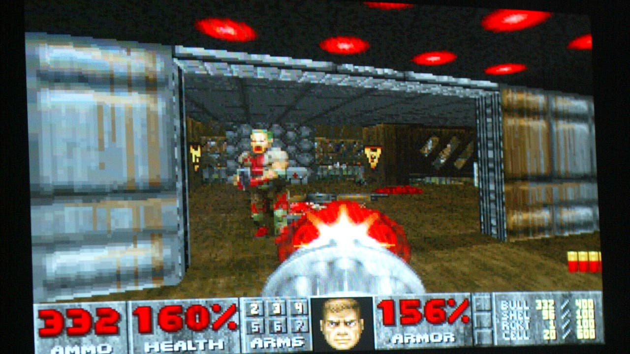 Ein Bildschirmfoto eines klassischen Egoshooter-Videospiels, das auf einem Monitor angezeigt wird. Im Vordergrund ist eine Hand auf einer Tastatur zu sehen, die das Spiel steuert. Auf dem Bildschirm ist ein Spielcharakter in einer futuristischen Umgebung zu erkennen, der eine Waffe abfeuert. Unten auf dem Bildschirm sind Statusanzeigen für Munition, Gesundheit und Rüstung zu sehen. Das Bild vermittelt eine aktive Spielszene.