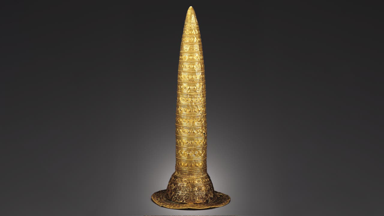 Ein konischer, goldener Hut mit detaillierten, kreisförmigen und linearen Mustern, die die Oberfläche bedecken. Der Hut hat eine breite Krempe und ist auf einem neutralen, dunklen Hintergrund abgebildet. Die Verzierungen sind symmetrisch und gleichmäßig verteilt, was auf eine kunstvolle Handwerkskunst hinweist.