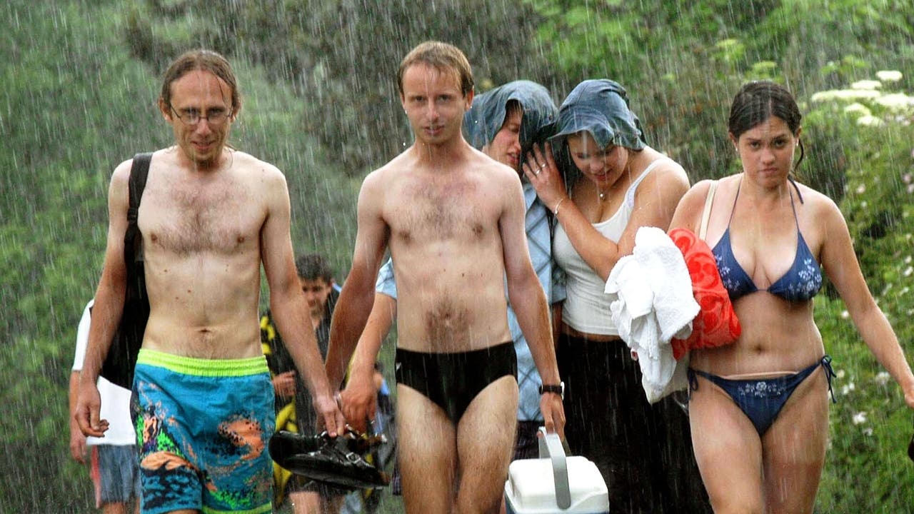 Eine Gruppe von fünf Menschen in Badebekleidung läuft im Regen einen Weg entlang. Zwei Männer tragen Badehosen, einer hält Schuhe in der Hand. Zwei Frauen schützen sich mit einem Handtuch über dem Kopf, während die dritte Frau ein Handtuch und Schuhe trägt. Im Hintergrund ist grüne Vegetation zu sehen. Die Stimmung wirkt unbeschwert trotz des Sommerregens.