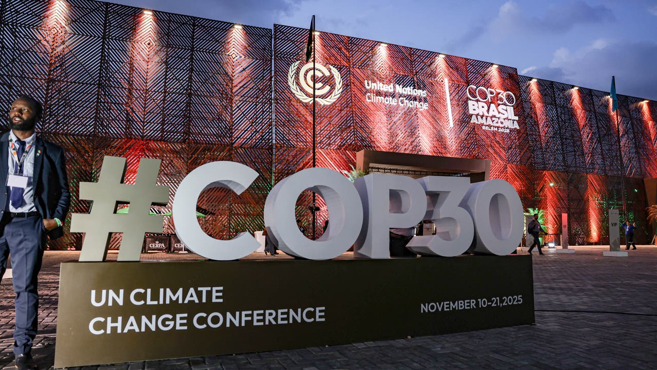 Eine große Skulptur mit der Aufschrift "#COP30" steht vor einem beleuchteten Gebäude, das für die UN-Klimakonferenz 2025 in Brasilien, Amazonas, vorbereitet ist. Auf dem Gebäude sind die Logos der Vereinten Nationen und der Konferenz sichtbar. Menschen gehen an der Szenerie vorbei. Der Himmel ist bewölkt, und die Umgebung ist in warmen Farben beleuchtet.