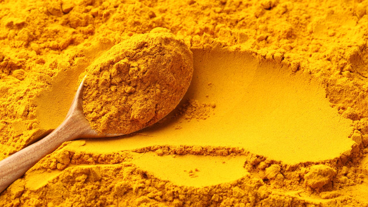 Ein Holzlöffel liegt auf einer Oberfläche aus leuchtend gelbem Pulver, das sich um den Löffel herum verteilt. Das Bild zeigt eine Nahaufnahme von Kurkumapulver, das für seine intensive Farbe und Verwendung in der Küche bekannt ist.