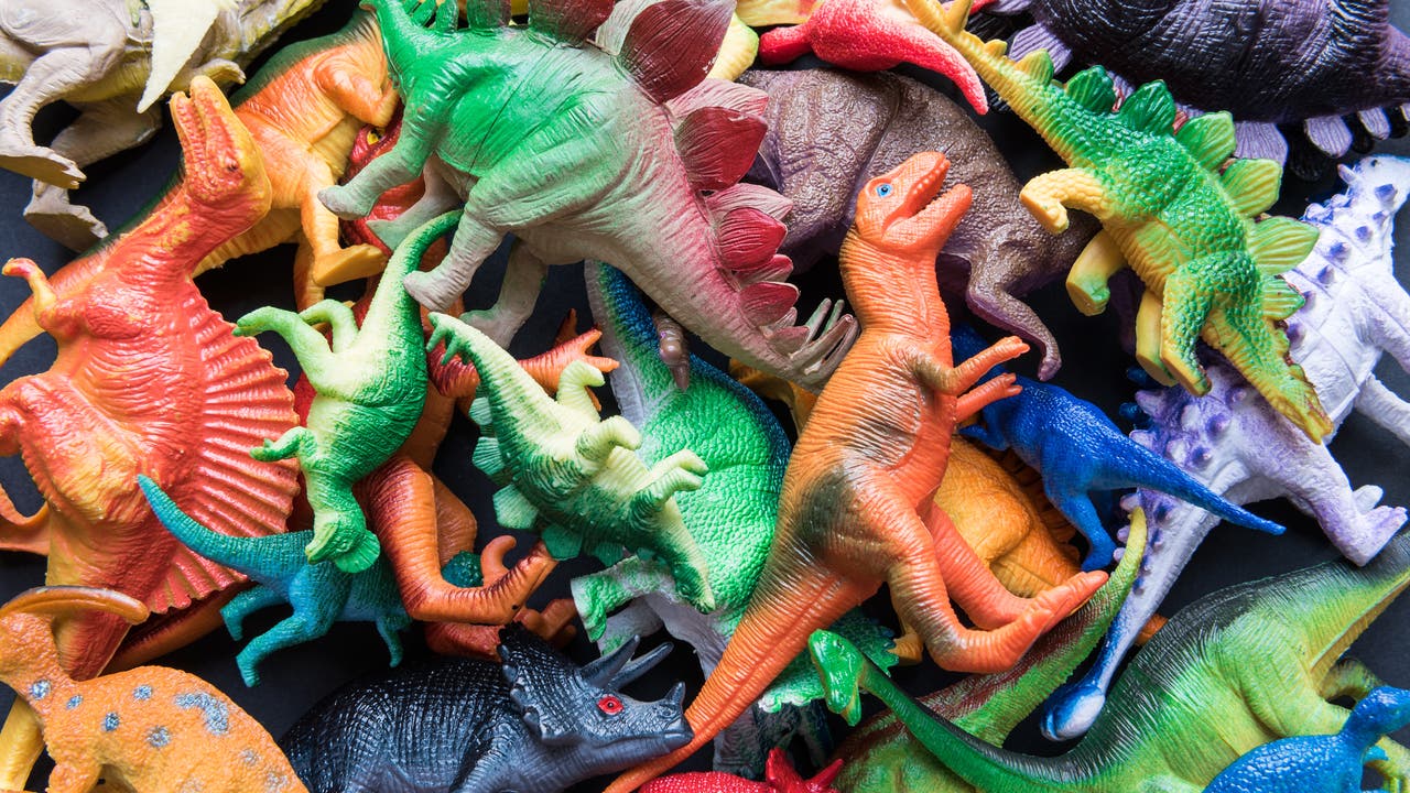 Eine Vielzahl bunter Dinosaurier-Spielzeugfiguren liegt dicht beieinander auf einer dunklen Oberfläche. Die Figuren sind in verschiedenen Farben wie Grün, Rot, Blau und Orange gestaltet und zeigen unterschiedliche Dinosaurierarten mit charakteristischen Merkmalen wie Hörnern, Platten und langen Schwänzen. Die Szene wirkt lebendig und chaotisch, da die Figuren in verschiedenen Richtungen ausgerichtet sind.