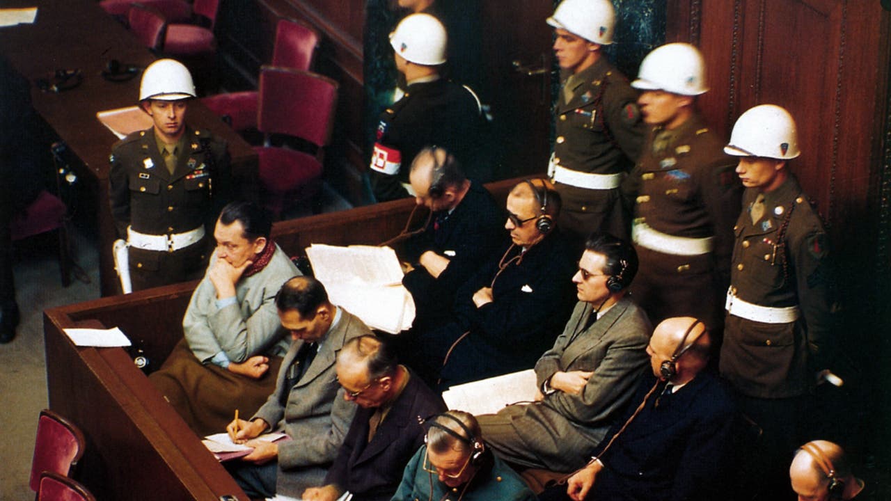 Eine Gruppe von Männern sitzt in einem Gerichtssaal, einige tragen Kopfhörer. Hinter ihnen stehen mehrere Soldaten in Uniform mit weißen Helmen. Die Szene vermittelt eine ernste und formelle Atmosphäre, typisch für ein Gerichtsverfahren. Die Männer scheinen aufmerksam zu sein, einige machen Notizen. Der Raum ist mit Holzwänden ausgestattet.