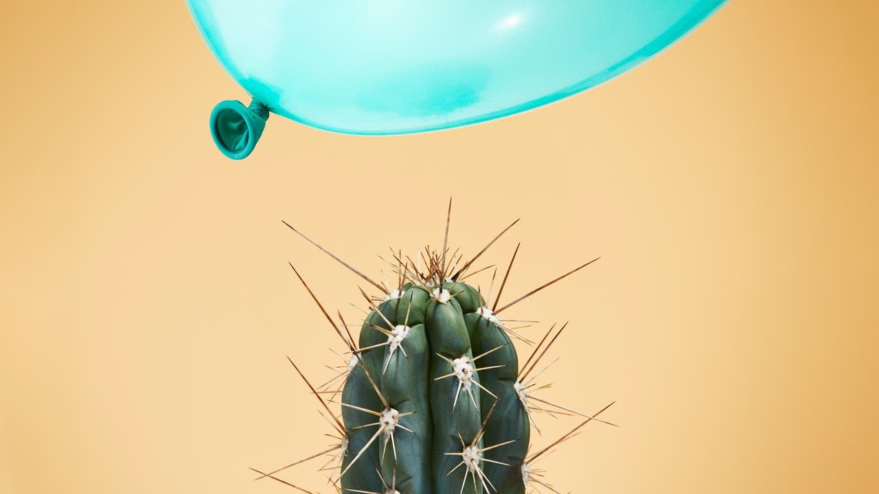 Ein grüner Kaktus mit langen, spitzen Stacheln steht vor einem gelben Hintergrund. Direkt über dem Kaktus schwebt ein hellblauer Luftballon, der kurz davor ist, die Stacheln zu berühren. Die Szene vermittelt Spannung und das Nahen eines Unglücks.