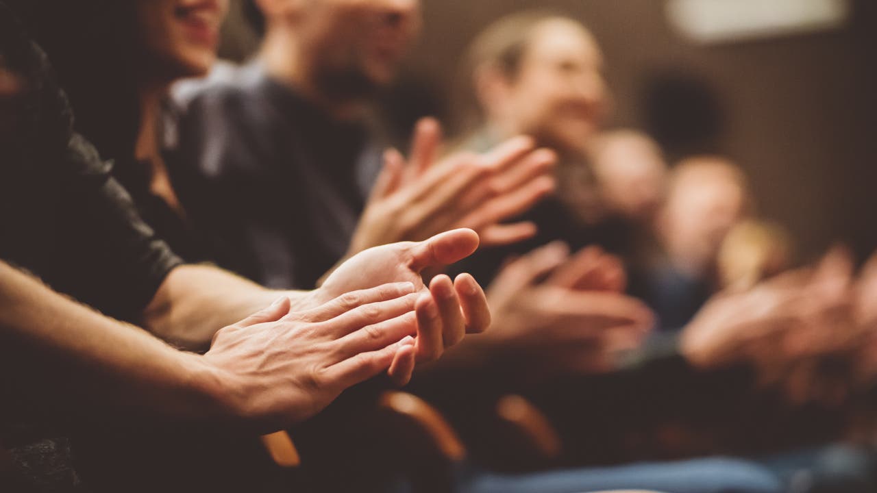 Eine Gruppe von Menschen sitzt in einem Auditorium und applaudiert. Die Hände im Vordergrund sind scharf gestellt, während die Gesichter im Hintergrund unscharf sind.