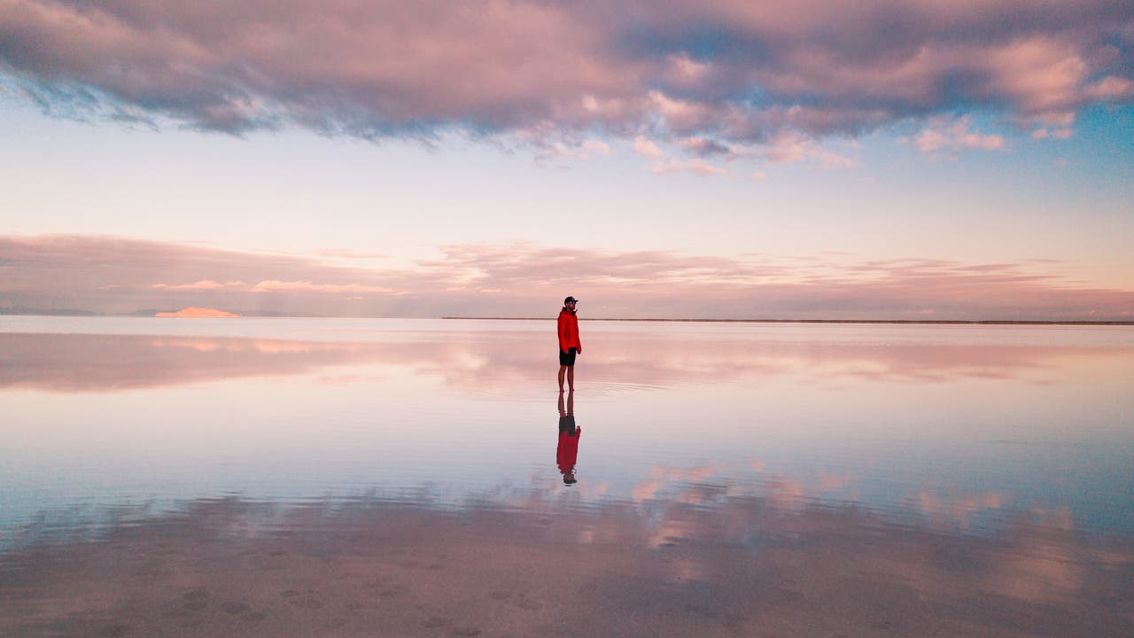Eine Person in einer roten Jacke steht inmitten einer glatten Wasseroberfläche, auf der sich der Himmel mit rosa und blauen Wolken spiegelt. Der Horizont ist in der Ferne sichtbar, während die Wolken den Himmel dramatisch gestalten. Es handelt sich hier um die überfluteten Salzebenen "Bonneville Salt Flats" in Utah, USA.