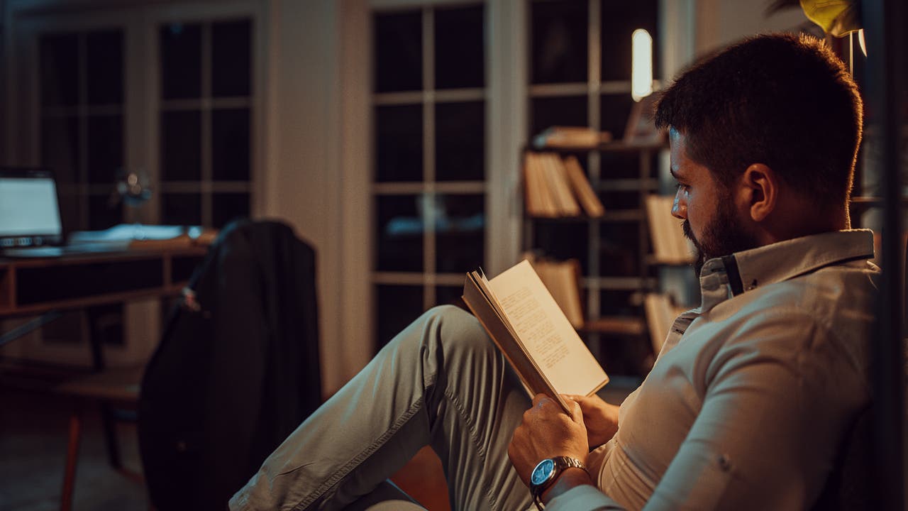 Ein Mann sitzt in einem gemütlichen Raum und liest ein Buch. Er trägt ein helles Hemd und eine Uhr am Handgelenk. Im Hintergrund sind ein Schreibtisch mit einem Laptop und ein Bücherregal zu sehen. Der Raum ist schwach beleuchtet, was eine entspannte Atmosphäre schafft.