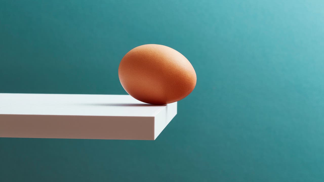 Ein braunes Ei balanciert am Rand einer weißen, wie ein Sprungbrett wirkenden Plattform vor einem türkisfarbenen Hintergrund. Das Bild vermittelt ein Gefühl von Spannung und Balance. Es gibt keinen Text oder eine URL im Bild.