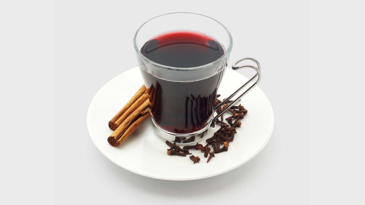 Ein Glas mit rotem Glühwein, umgeben von Zimtstangen und Nelken, auf einem grauen Hintergrund. Das Glas hat einen Metallgriff. 