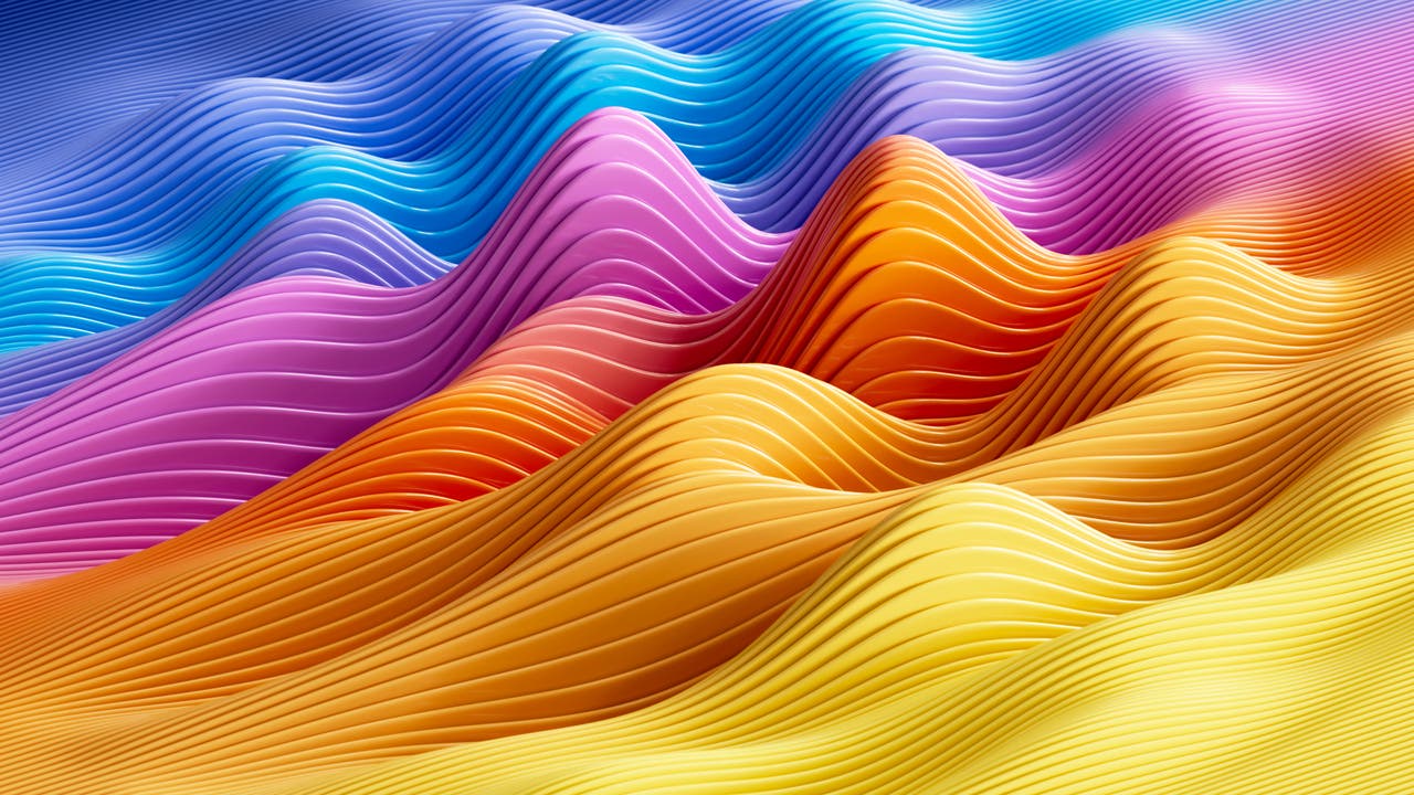 Abstrakte Darstellung wellenförmiger Strukturen in lebendigen Farben. Die Wellen verlaufen von links nach rechts und zeigen eine Farbpalette von Blau über Lila, Pink, Orange bis Gelb. Jede Welle besteht aus feinen, parallelen Linien, die eine dynamische Bewegung suggerieren. Der Hintergrund ist in einem sanften Blau gehalten, das in den oberen Bereich des Bildes übergeht. Die Komposition vermittelt ein Gefühl von Energie und Fluss.