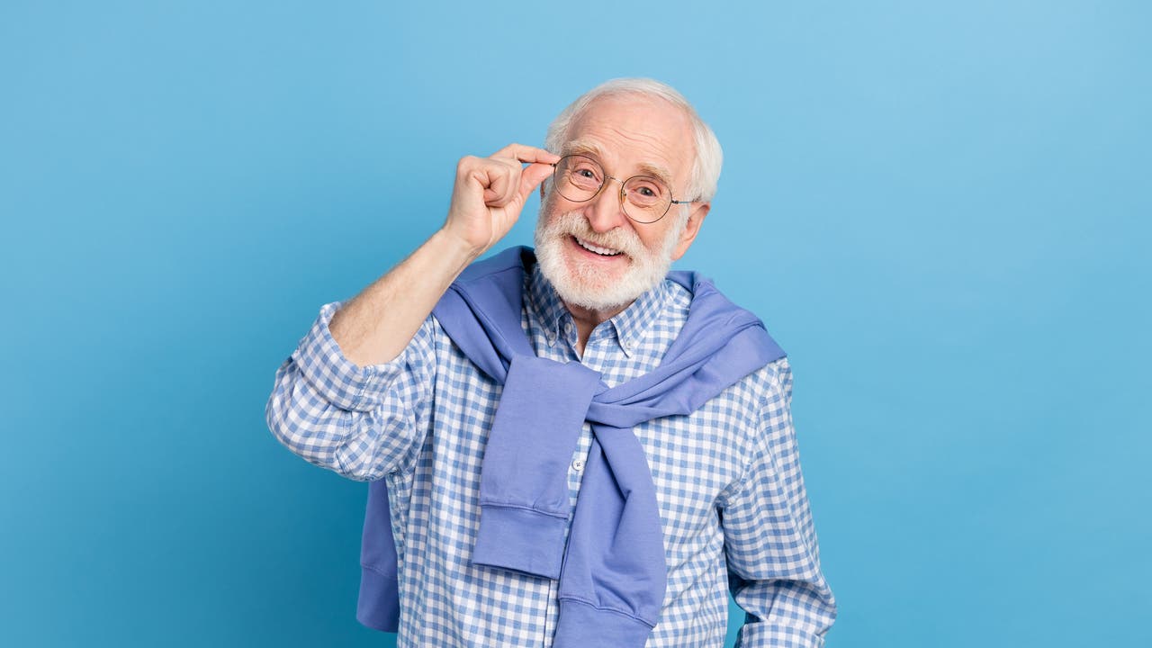 Ein älterer Mann mit Brille lächelt freundlich in die Kamera. Er trägt ein kariertes Hemd und hat einen Pullover um die Schultern gebunden. Der Hintergrund ist einfarbig blau.