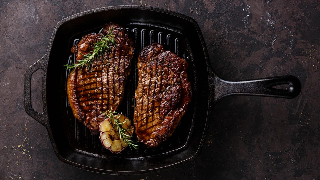 Foto von zwei kross gebratenen Rindersteaks in einer eckigen, gusseisernen Pfanne, garniert mit Rosmarinzweigen