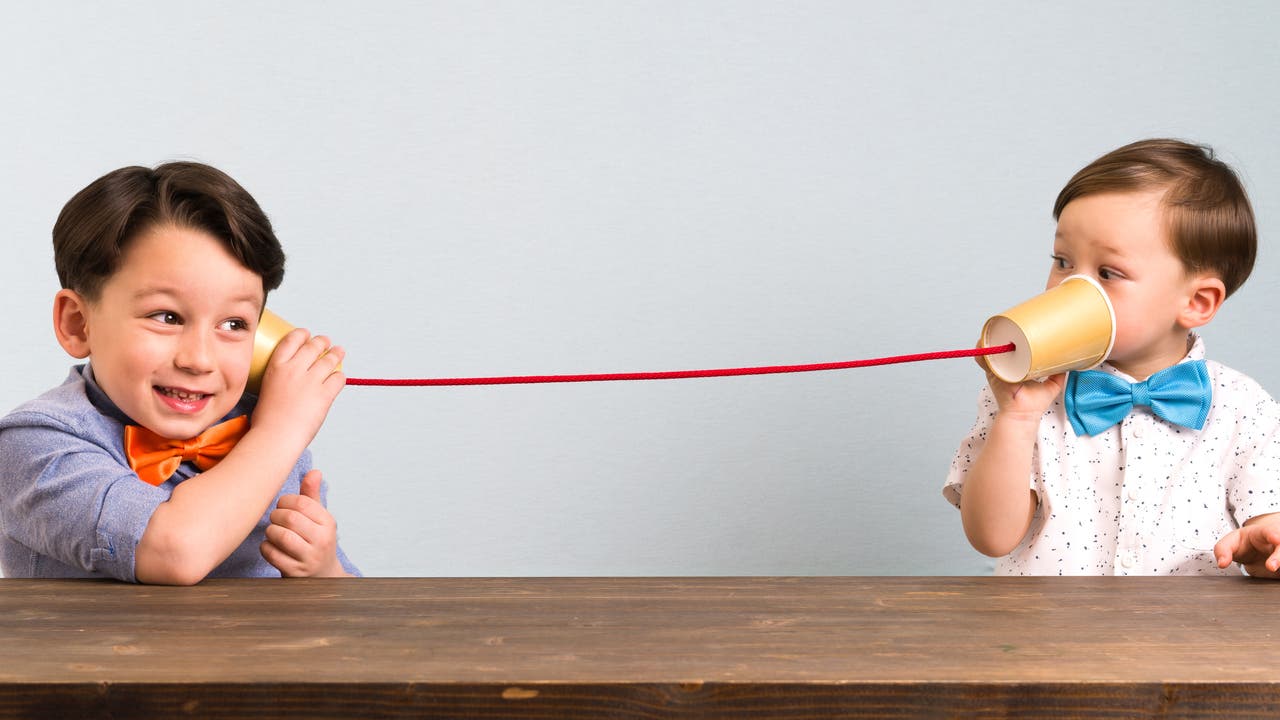 Zwei Kinder spielen mit einem selbst gemachten Telefon aus Pappbechern und einer Schnur. Das linke Kind hält einen Becher ans Ohr und lächelt, während das rechte Kind in den anderen Becher spricht. Beide tragen bunte Fliegen. Sie sitzen an einem Holztisch vor einem neutralen Hintergrund.
