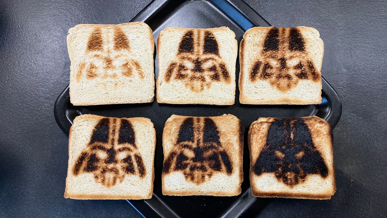 Sechs Scheiben Toastbrot auf einem schwarzen Tablett, jede mit einem dunklen, gebräunten Muster, das einem ikonischen Helm ähnelt. Links unten ist eine kleine, schwarze Tasse zu sehen. Der Hintergrund ist eine dunkle Oberfläche.