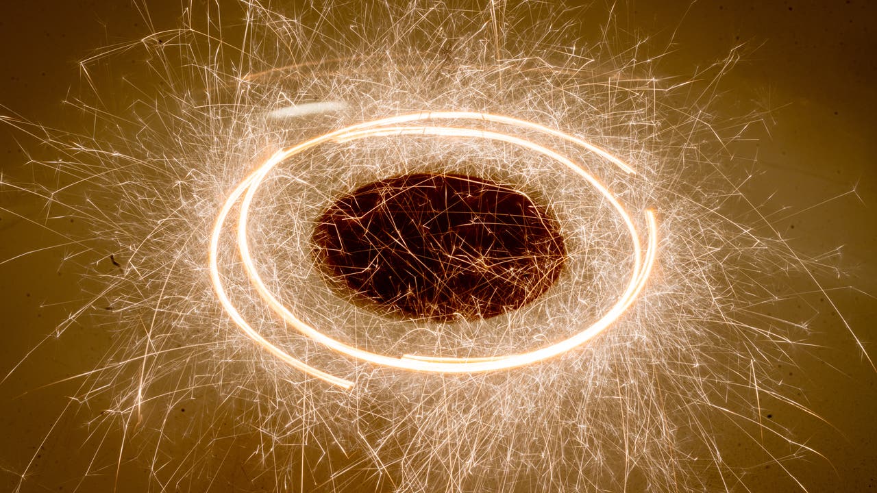 Brennende Wunderkerzen, die tangential an einer Scheibe aufgehängt sind, hinterlassen sprühende Funken, während sich die Scheibe im Kreis dreht. Eine lange Belichtungsdauer macht die glühenden Stäbe als Strichspuren sichtbar.