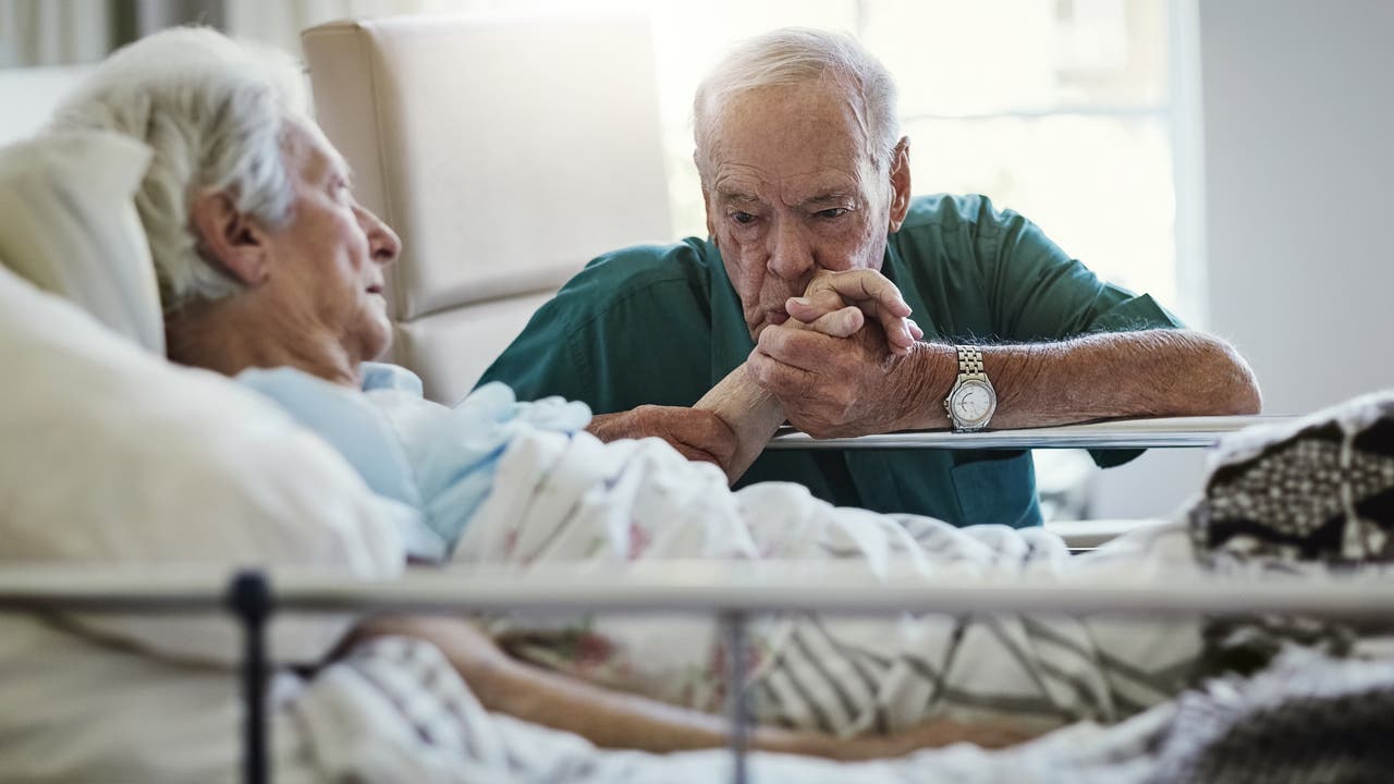 Ein Mann sitzt am Krankenbett seiner Frau und küsst zärtlich ihre Hand.