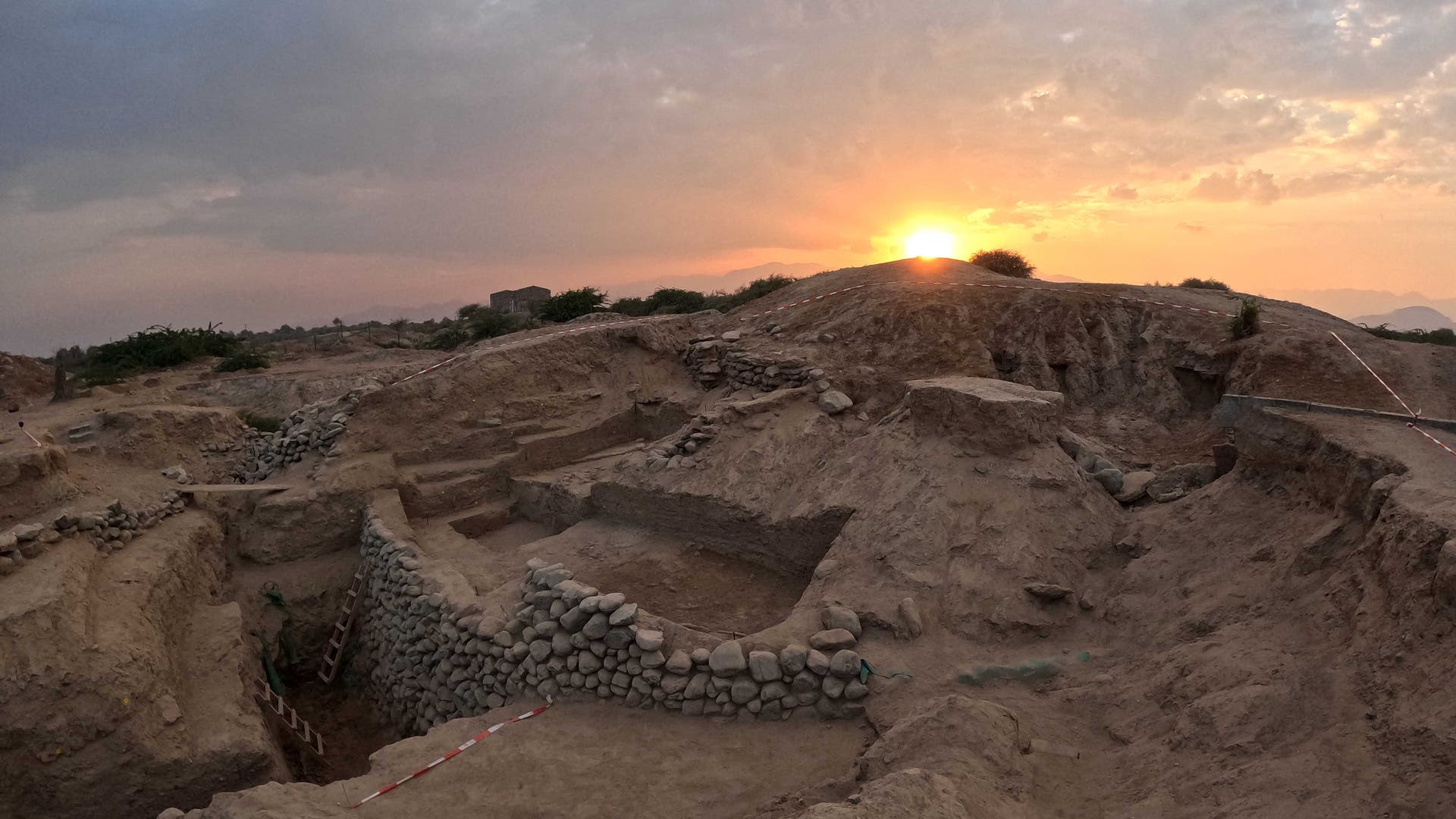 Ein Erdhügel mit frei gelegten Steinmauern: Die Ausgrabungsstätte Kalba in den Vereinigten Arabischen Emiraten während der Abenddämmerung.