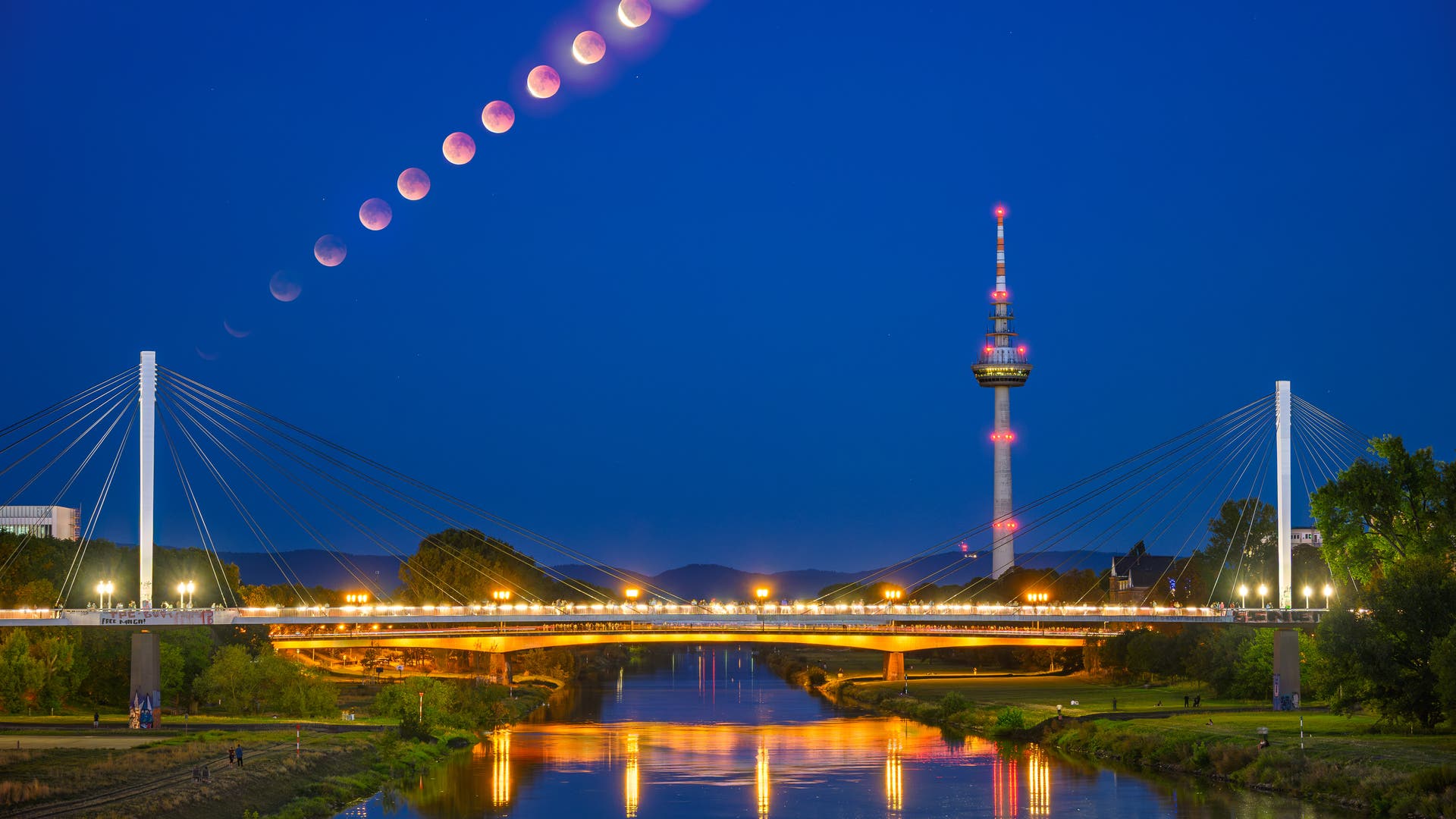Eine nächtliche Stadtlandschaft mit einer beleuchteten Brücke und einem Fernsehturm im Vordergrund. Am Himmel ist eine Abfolge von Mondphasen während einer Mondfinsternis zu sehen, die von links nach rechts verläuft. Der Fluss unter der Brücke spiegelt die Lichter wider, und der Himmel ist klar und dunkelblau. Schlüsselwörter: Mondfinsternis, Brücke, Fernsehturm, Nacht, Reflexion.