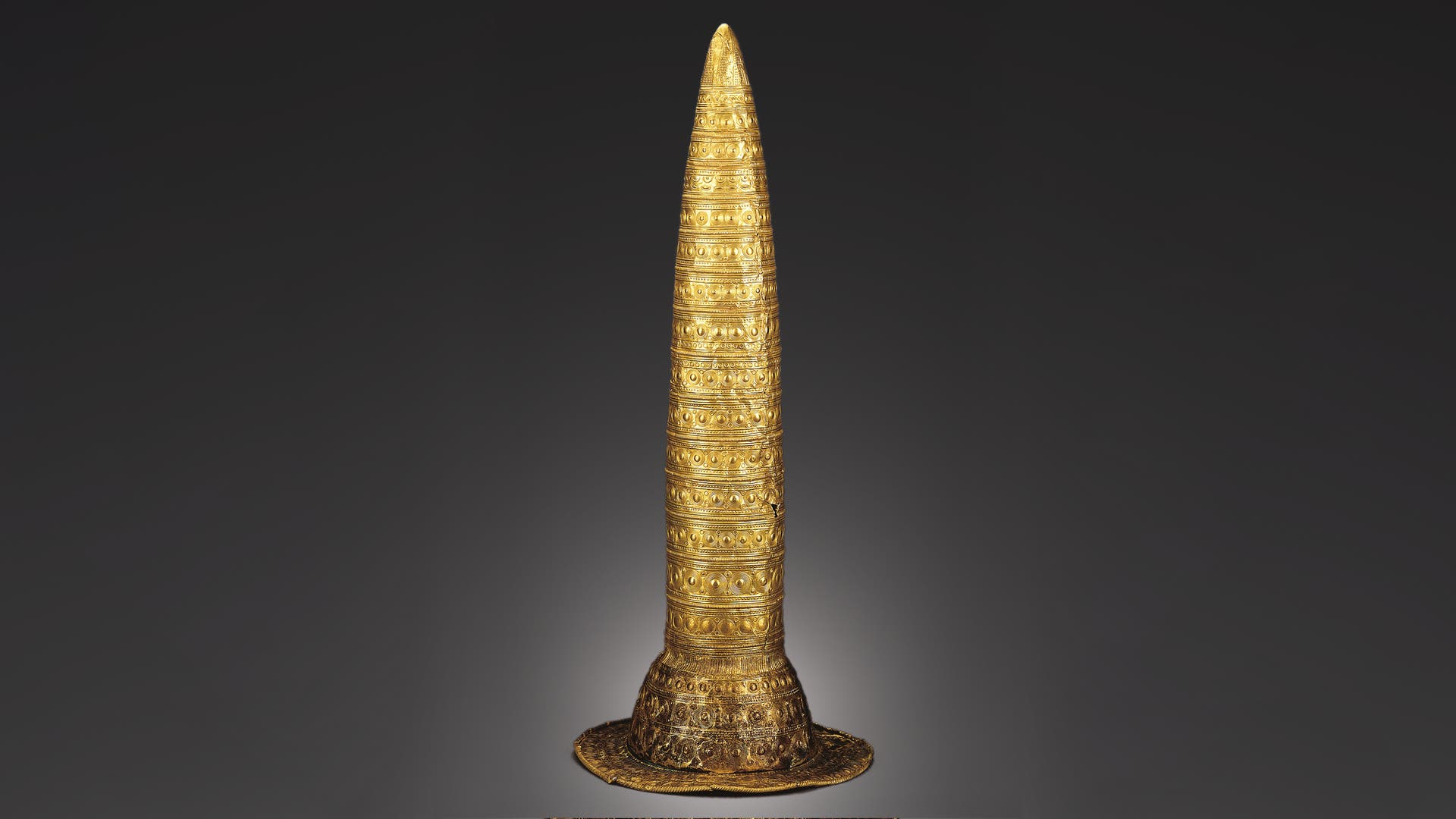 Ein konischer, goldener Hut mit detaillierten, kreisförmigen und linearen Mustern, die die Oberfläche bedecken. Der Hut hat eine breite Krempe und ist auf einem neutralen, dunklen Hintergrund abgebildet. Die Verzierungen sind symmetrisch und gleichmäßig verteilt, was auf eine kunstvolle Handwerkskunst hinweist.