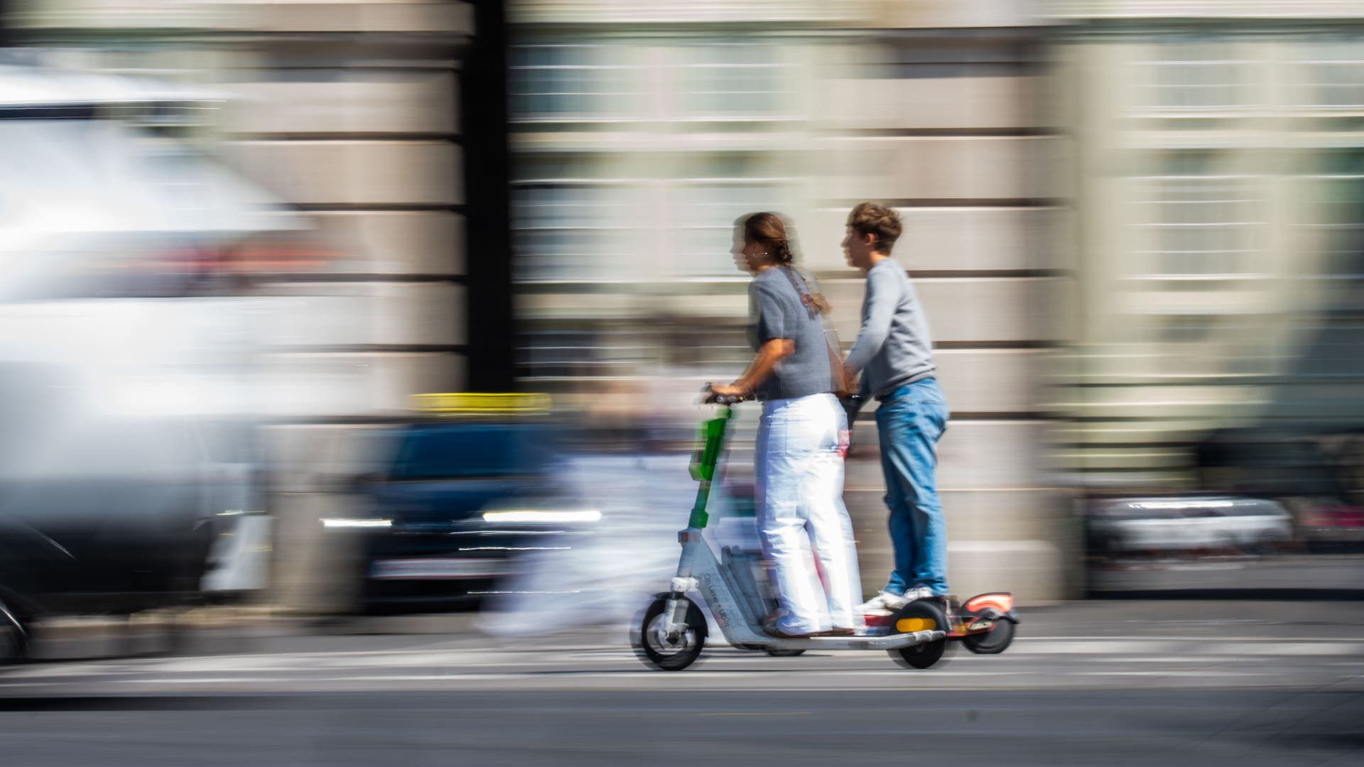 Zwei Personen fahren gemeinsam auf einem E-Scooter durch eine belebte Stadtstraße. Die Umgebung ist verschwommen, was die Bewegung und Geschwindigkeit betont. Im Hintergrund sind Gebäude und vorbeifahrende Autos zu erkennen.