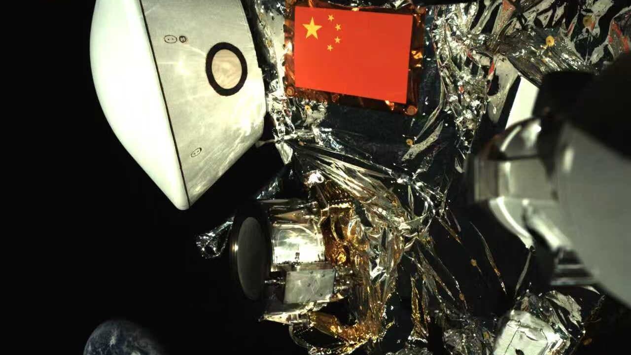 Eine Raumsonde im Weltraum, umgeben von reflektierenden Materialien und technischen Komponenten. Eine chinesische Flagge ist prominent auf der Sonde zu sehen. Im Hintergrund ist die Erde als kleiner blauer Planet sichtbar. Das Bild zeigt die Komplexität und den technologischen Fortschritt der Raumfahrt. Unten links befinden sich Logos, die auf chinesische Raumfahrtorganisationen hinweisen.