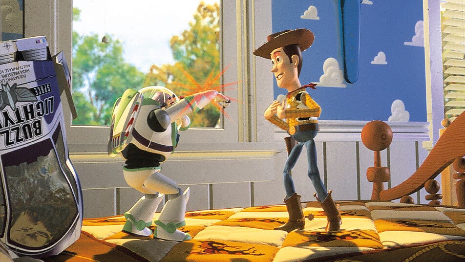 Eine animierte Szene zeigt zwei Spielzeugfiguren in einem Kinderzimmer. Links steht eine Figur in einem Raumanzug, die mit einem ausgestreckten Arm einen roten Laserstrahl abfeuert. Rechts steht eine Cowboy-Figur mit einem Hut, die lächelnd zuschaut. Im Hintergrund ist ein Fenster mit Blick auf Bäume und ein blauer Himmel mit Wolkenmuster an der Wand zu sehen. Auf dem Bett liegt eine bunte Decke mit Cowboy-Motiven. Links im Bild ist eine Verpackung mit der Aufschrift "Buzz Lightyear" zu erkennen.