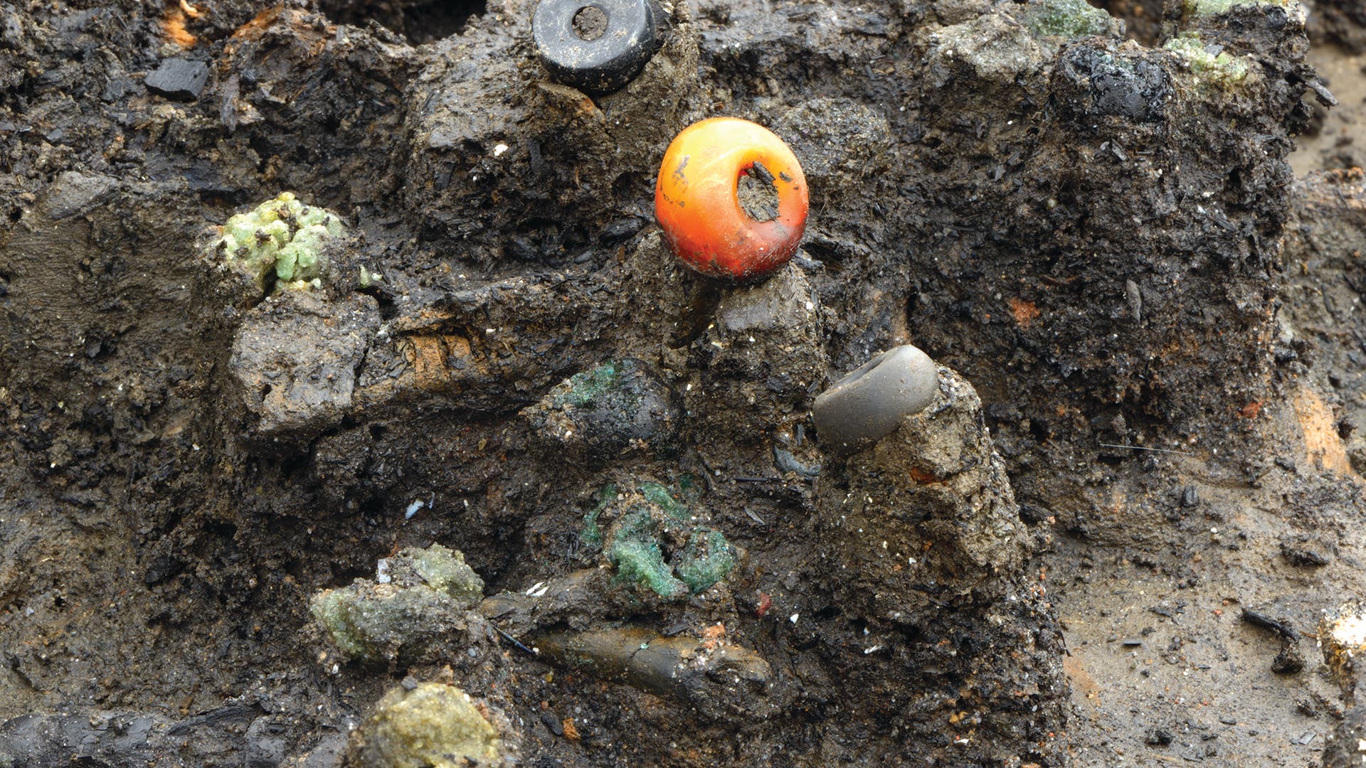 Eine Nahaufnahme von schlammigem Boden, in dem mehrere bunte Perlen eingebettet sind. Eine auffällige Ringperle aus Bernstein befindet sich in der Mitte, umgeben von schwarzen und grünen Glasperlen. Die Oberfläche ist uneben und zeigt Spuren von Feuchtigkeit und Verwitterung.