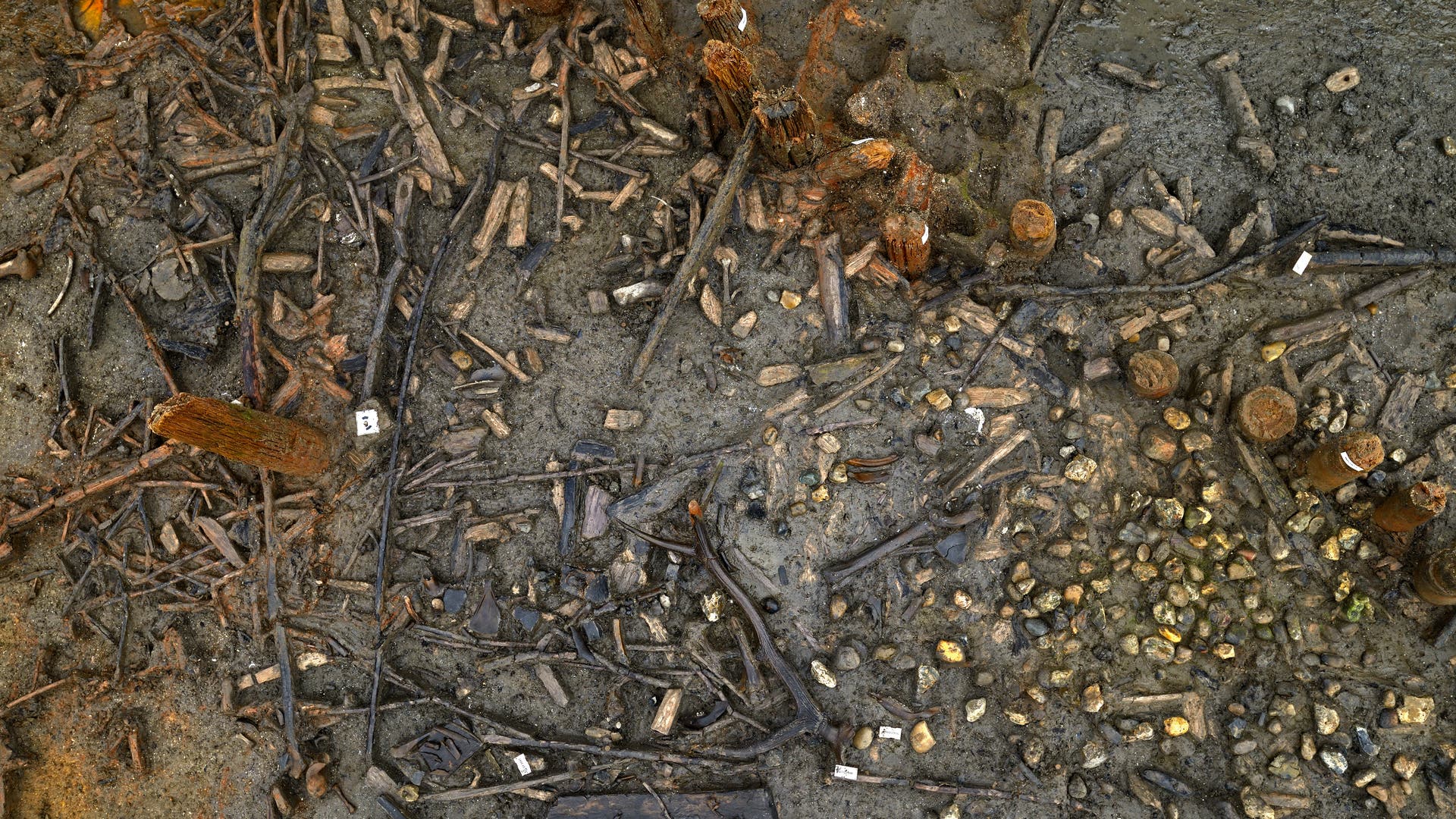 Eine archäologische Ausgrabungsstätte von oben mit freigelegten Holzstrukturen und kleinen nummerierten Markierungen auf dem Boden. Die Szene zeigt eine Vielzahl von Holzstücken und Steinen, die in einem erdigen, feuchten Bereich verteilt sind. Die Markierungen scheinen zur Kategorisierung oder Dokumentation der Fundstücke zu dienen.