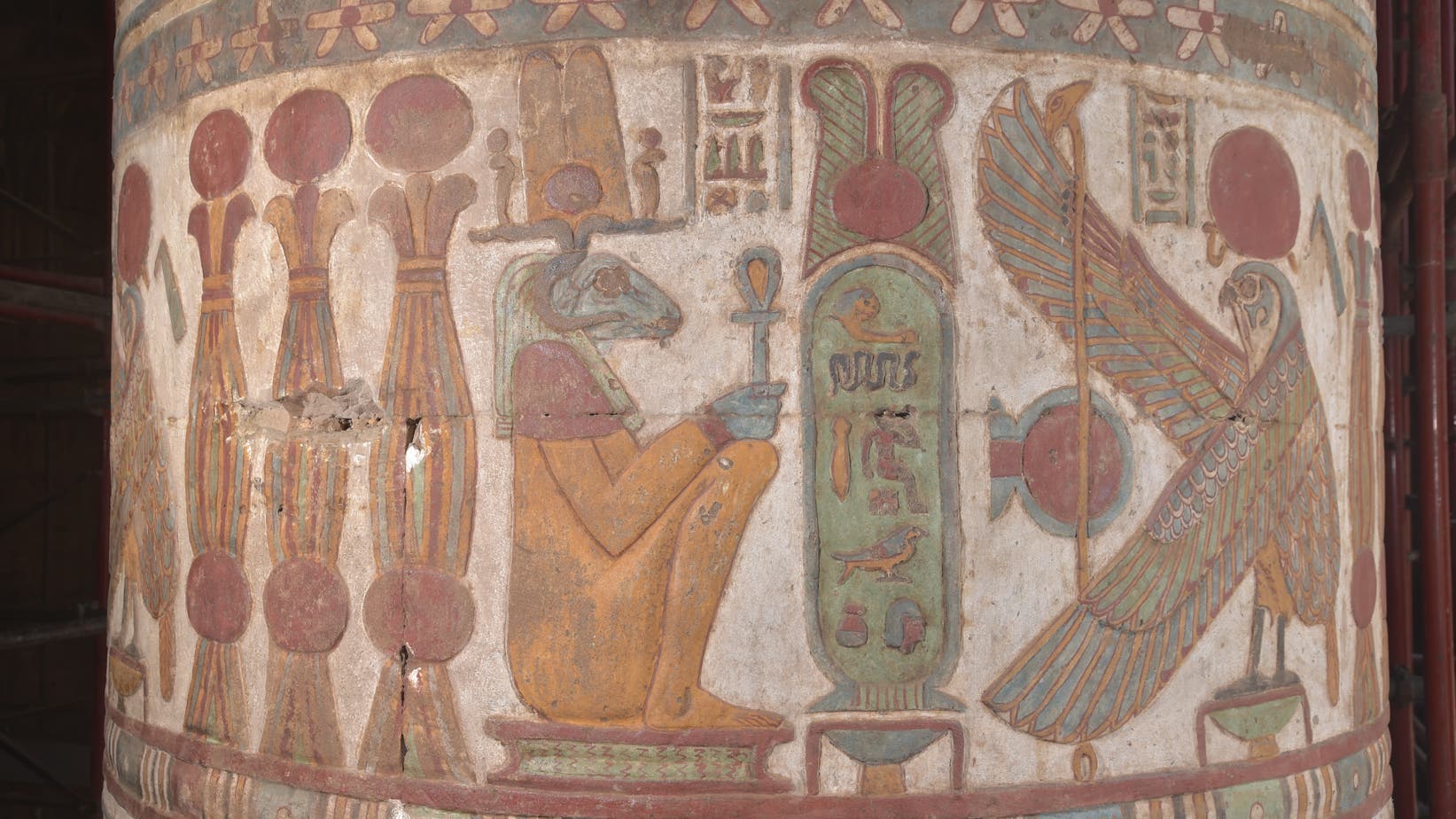 Eine antike ägyptische Säule mit farbenfrohen Hieroglyphen und Darstellungen. Links sind drei stilisierte Säulen zu sehen. Rechts davon hockt eine Figur mit einem Widderkopf, die ein Anch-Zepter hält. In der Mitte ist eine grün ausgemalte Kartusche mit Krone zu erkennen. Rechts ist ein großer, detaillierter Falke mit ausgebreiteten Flügeln abgebildet. Die Säule ist mit einem Band aus Blumenmustern verziert. Die Farben sind gut erhalten und zeigen Rot-, Blau-, Grün- und Gelbtöne.