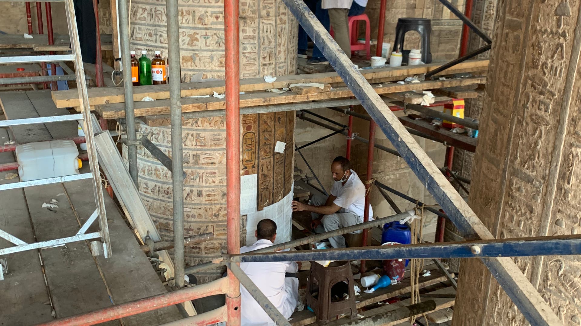 Mehrere Personen in weißen Kitteln arbeiten an der Restaurierung antiker, mit Hieroglyphen verzierter Säulen in einem historischen Gebäude. Sie sind von einem Gerüst umgeben, das den Zugang zu den hohen Strukturen ermöglicht. Die Szene ist gut beleuchtet, und verschiedene Restaurierungswerkzeuge sind sichtbar.