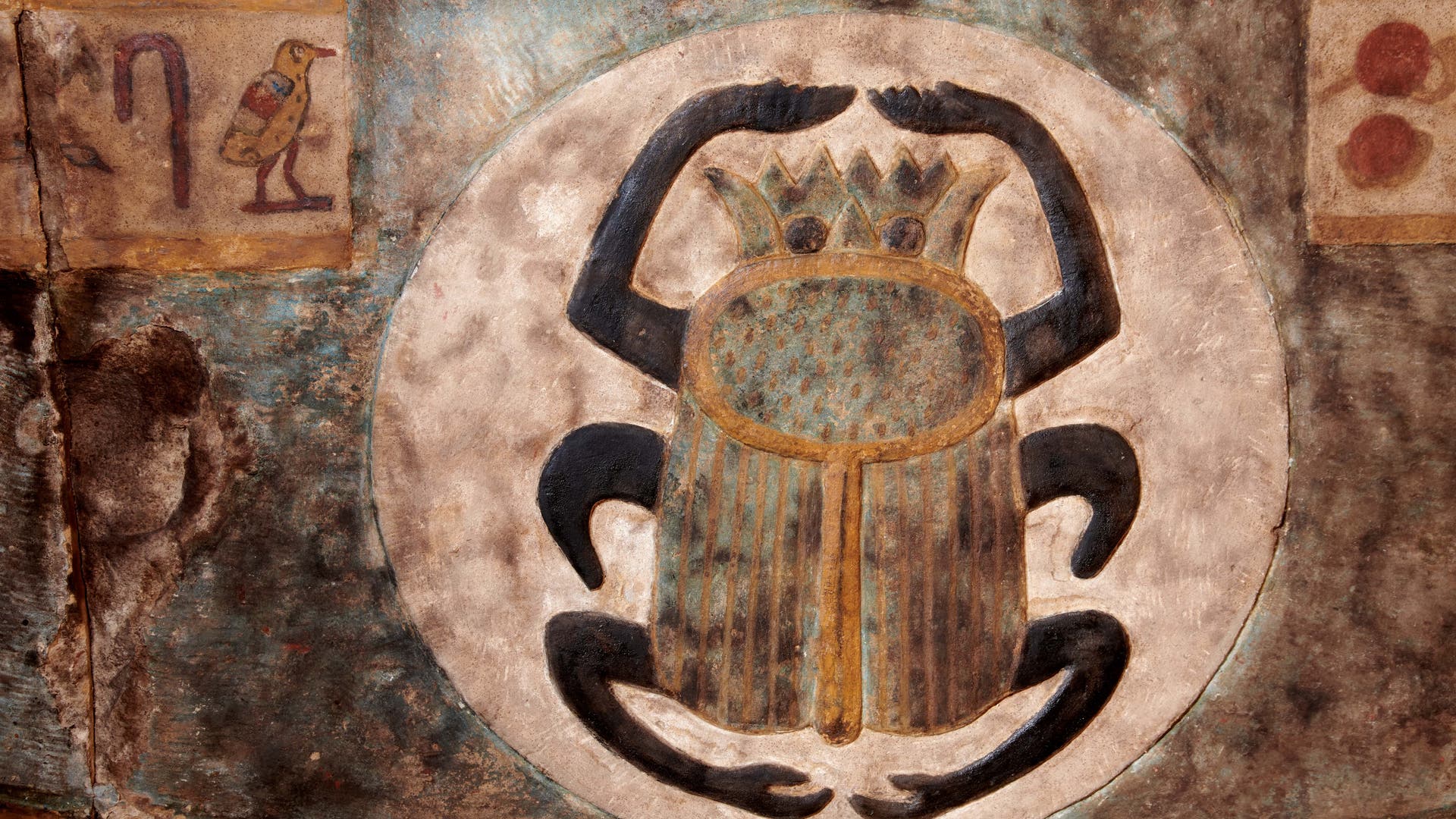 Ein antikes ägyptisches Wandbild zeigt einen Skarabäus, der in einem hellen Kreis dargestellt ist. Der Käfer hat detaillierte Flügel und eine zackenförmigen Kopf. Links ist ein Hieroglyphensymbol mit einem Vogel zu sehen. Rechts sind zwei weitere Hieroglyphen mit roten Sonnenscheiben abgebildet. Die Farben sind überwiegend erdige Töne mit blauen und goldenen Akzenten. 