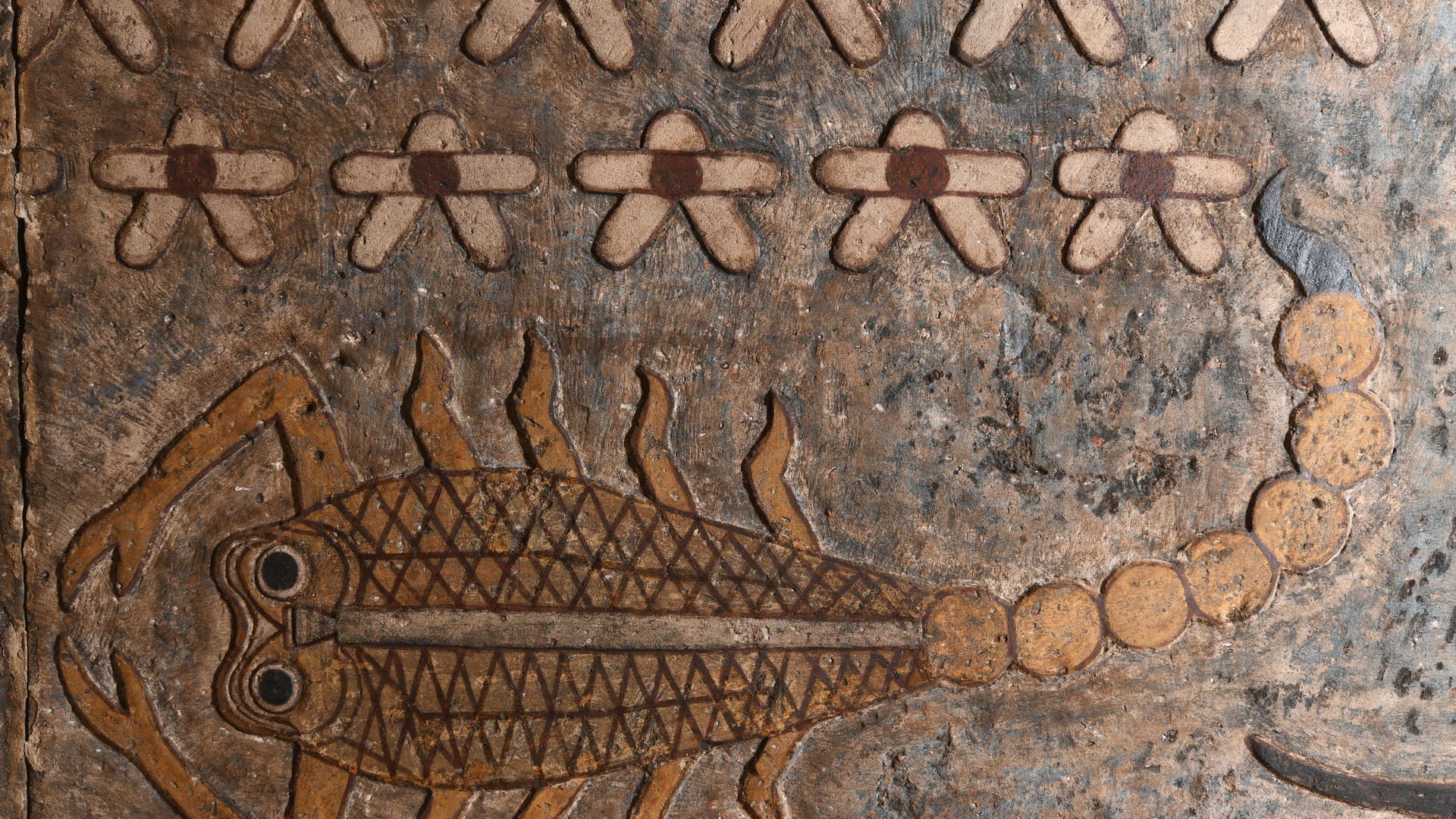 Ein antikes ägyptisches Relief zeigt einen stilisierten Skorpion mit detailliertem Körpermuster und großen Augen. Über dem Skorpion sind zwei Reihen von stilisierten Sternen abgebildet. Die Farben sind erdige Töne mit einem verwitterten Aussehen. 