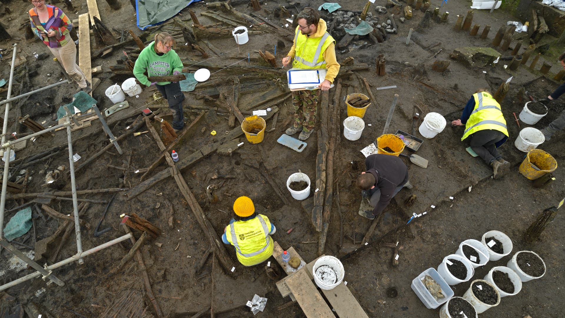 Eine Gruppe von Archäologen arbeitet auf einer Ausgrabungsstätte. Mehrere Personen in gelben Sicherheitswesten und einer in einem grünen Pullover sind zu sehen, während sie den Boden untersuchen und Proben in Eimern sammeln. Einer der Archäologen hält ein Klemmbrett. Die Umgebung ist mit Holzbalken und verschiedenen Werkzeugen übersät. Im Hintergrund sind Planen zu sehen, die den Bereich abdecken.