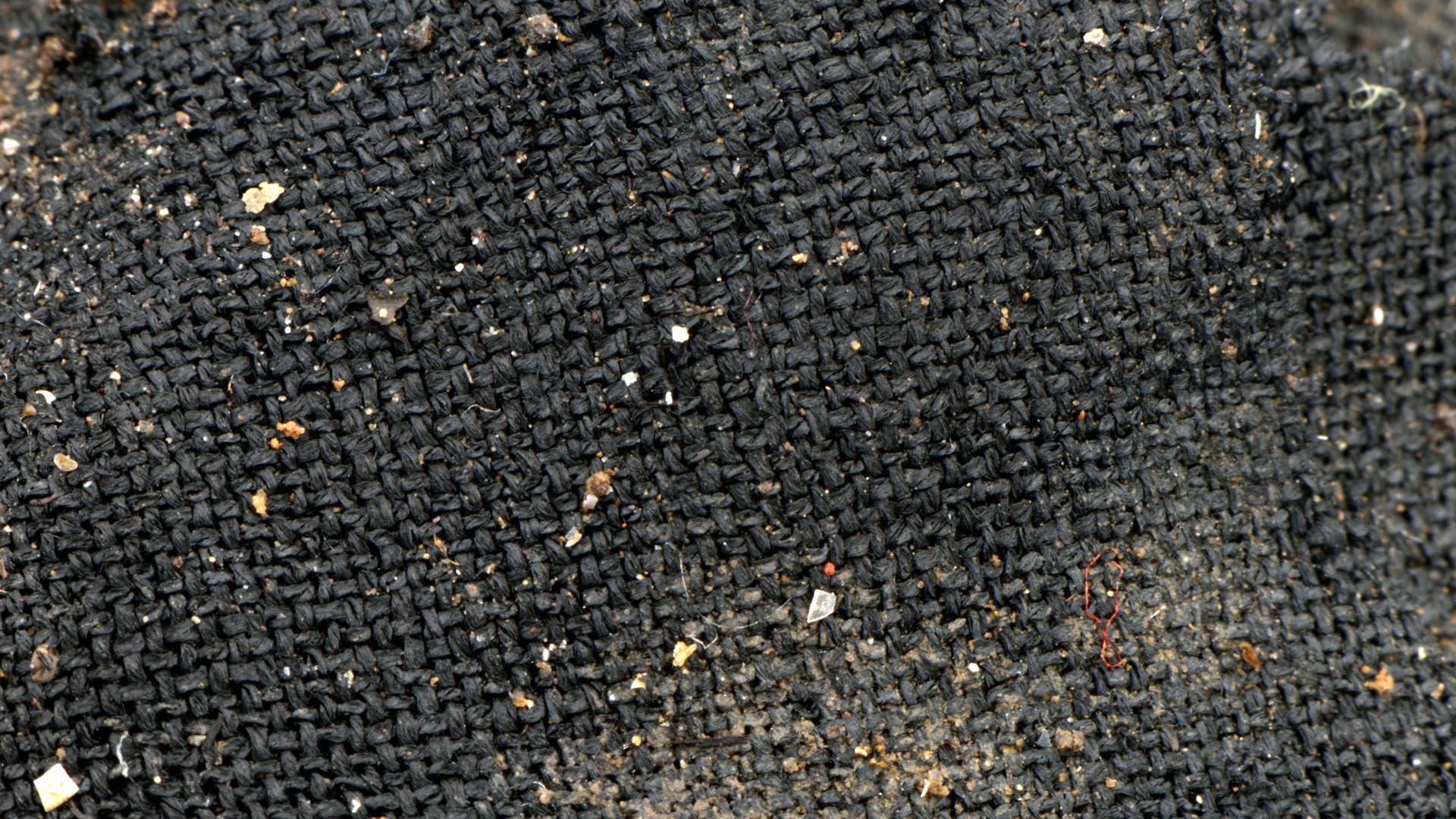 Nahaufnahme eines grob gewebten, schwarzen Stoffes mit sichtbaren Staub- und Schmutzpartikeln. Die Textur des Materials ist deutlich erkennbar, mit einem unregelmäßigen Muster aus kleinen Quadraten. Keine Menschen oder Text im Bild.