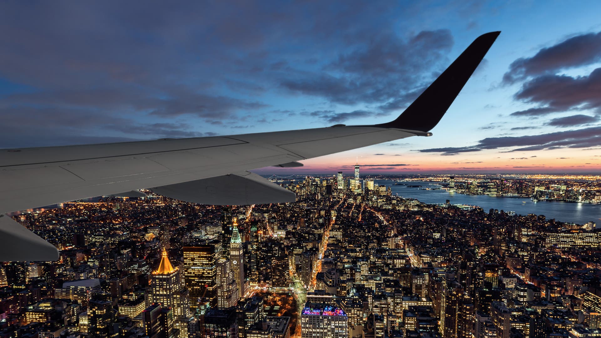 Blick aus einem Flugzeugfenster auf eine beleuchtete Stadtlandschaft bei Sonnenuntergang. Die Flügelspitze des Flugzeugs ist im Vordergrund sichtbar, während sich darunter ein dichtes Netz aus erleuchteten Straßen und Gebäuden erstreckt. Der Himmel zeigt eine Mischung aus Blau- und Orangetönen, und ein Fluss ist am Horizont zu erkennen.