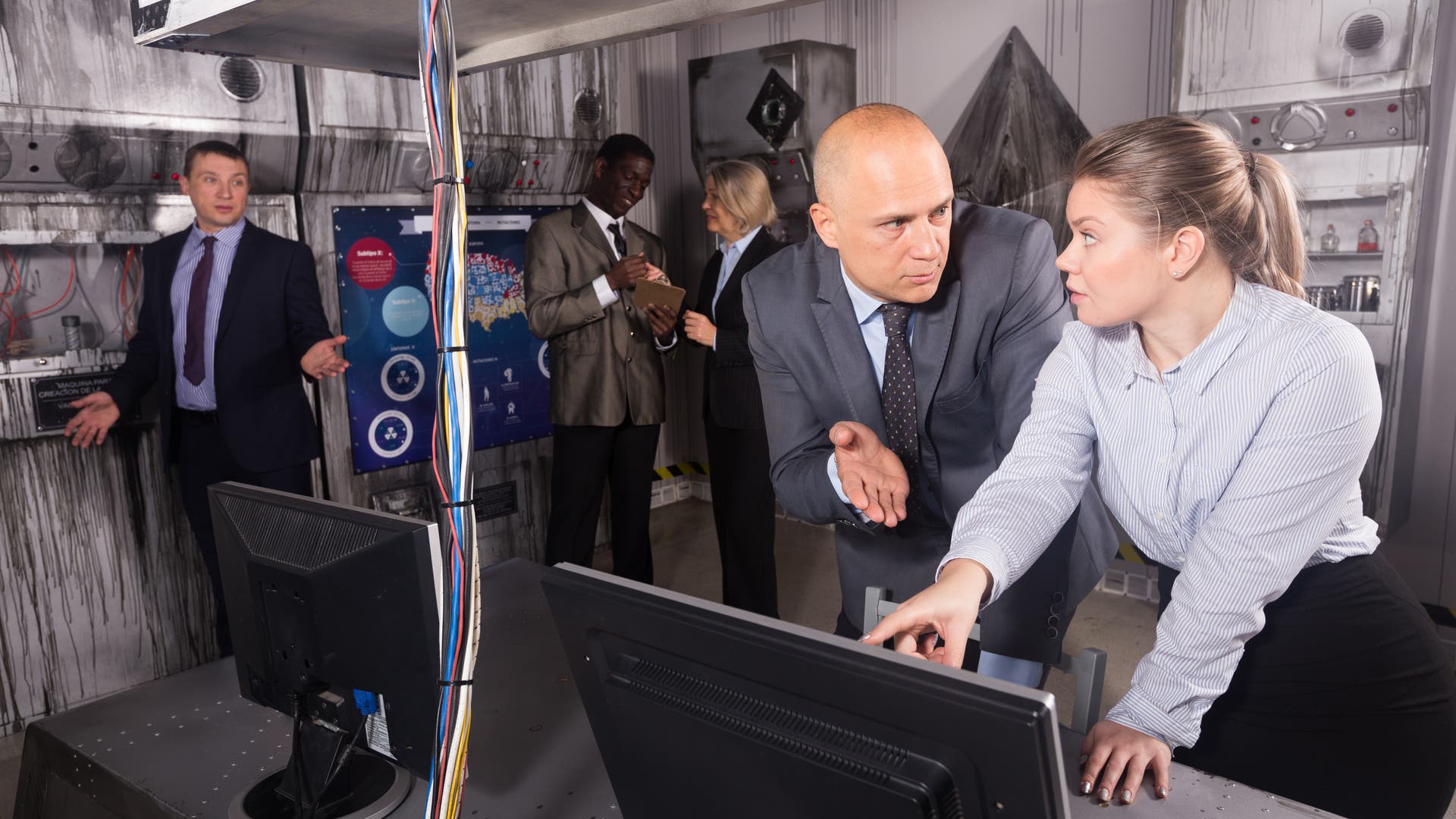 Eine Gruppe von Personen in einem Raum, der wie ein Kontrollzentrum aussieht. Zwei Personen im Vordergrund diskutieren angeregt über einen Computerbildschirm. Im Hintergrund sind weitere Personen zu sehen, die miteinander sprechen und Notizen machen. An den Wänden befinden sich technische Geräte und ein Poster mit Diagrammen. Die Szene vermittelt eine geschäftige und konzentrierte Arbeitsatmosphäre.