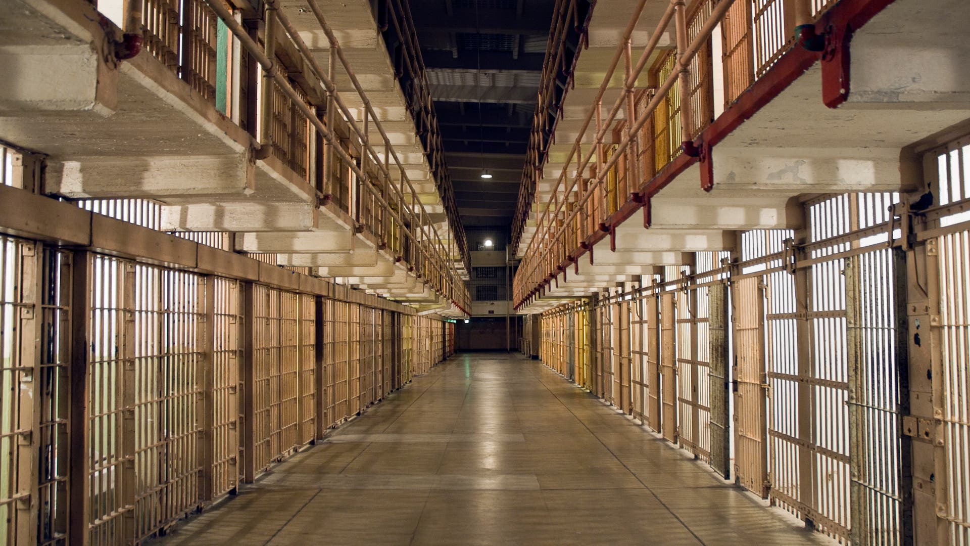 Ein langer, leerer Gefängnisflur mit Zellenreihen auf beiden Seiten. Die Zellen sind durch Gitterstäbe verschlossen, und das Licht fällt durch die Gitter, wodurch ein Muster auf dem Boden entsteht. Die Perspektive des Fotos betont die Tiefe des Flurs.